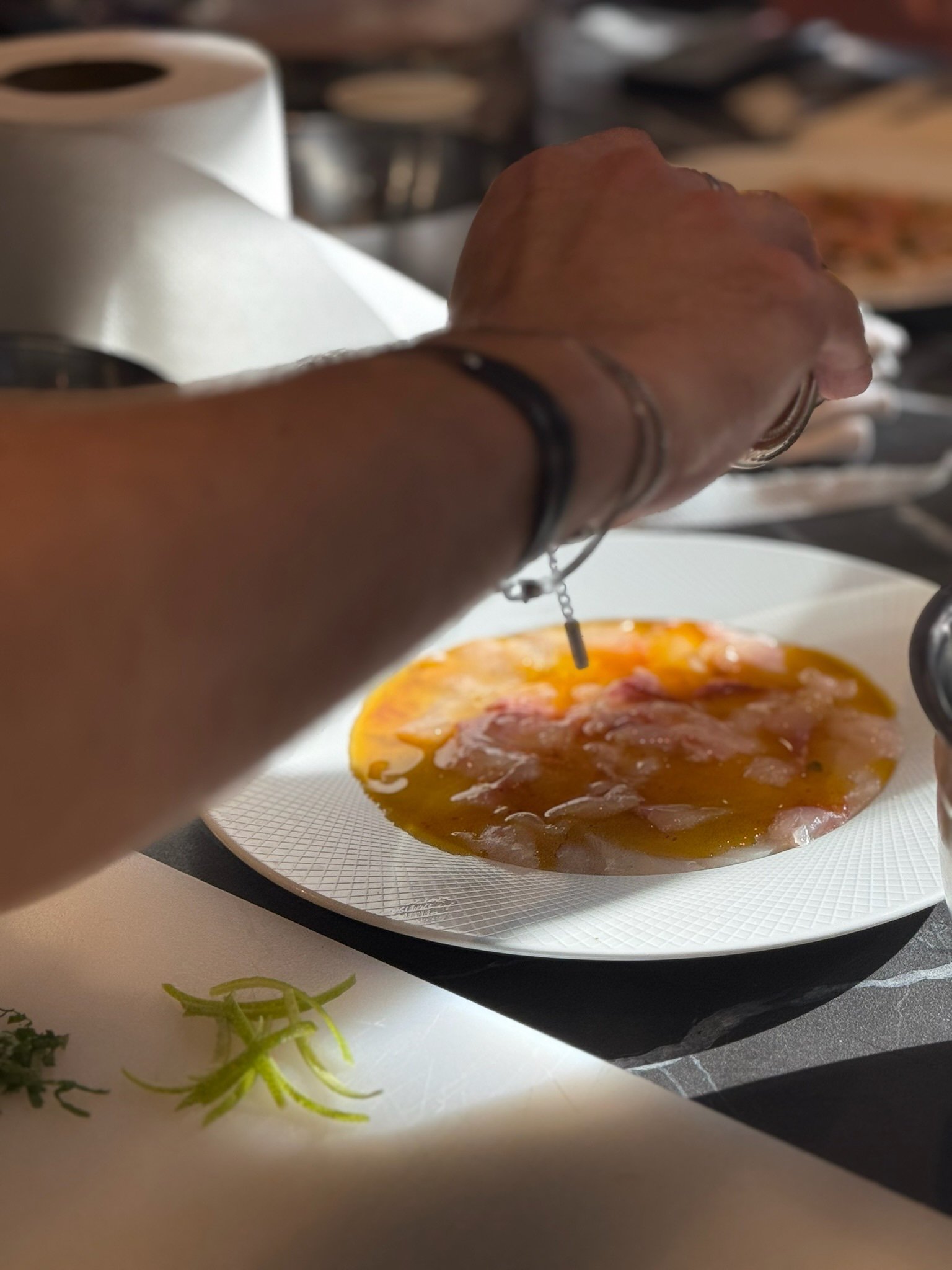 Main d'une personne assaisonnant du poisson cru tranché sur une assiette blanche.