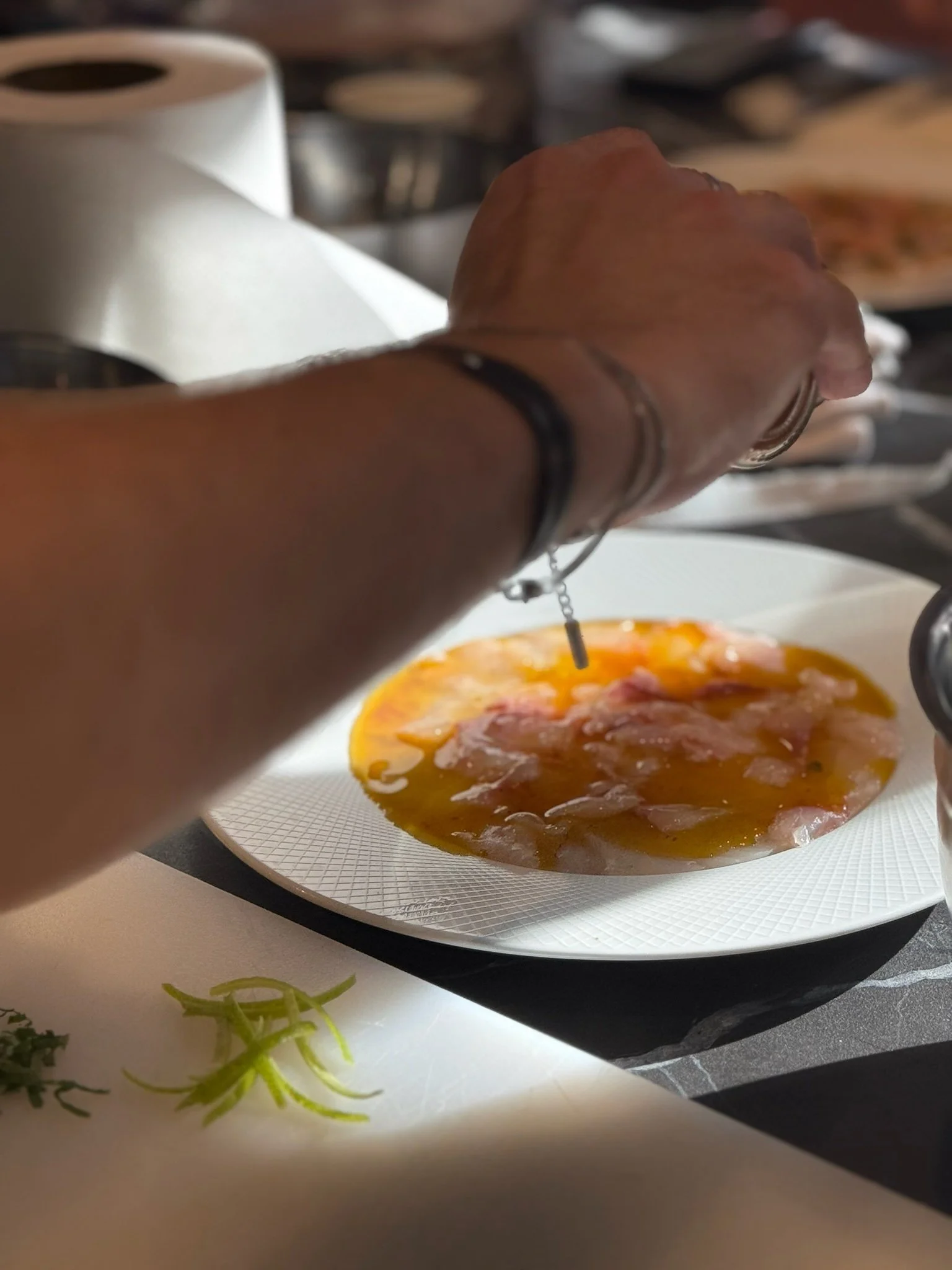 Main d'une personne assaisonnant du poisson cru tranché sur une assiette blanche.