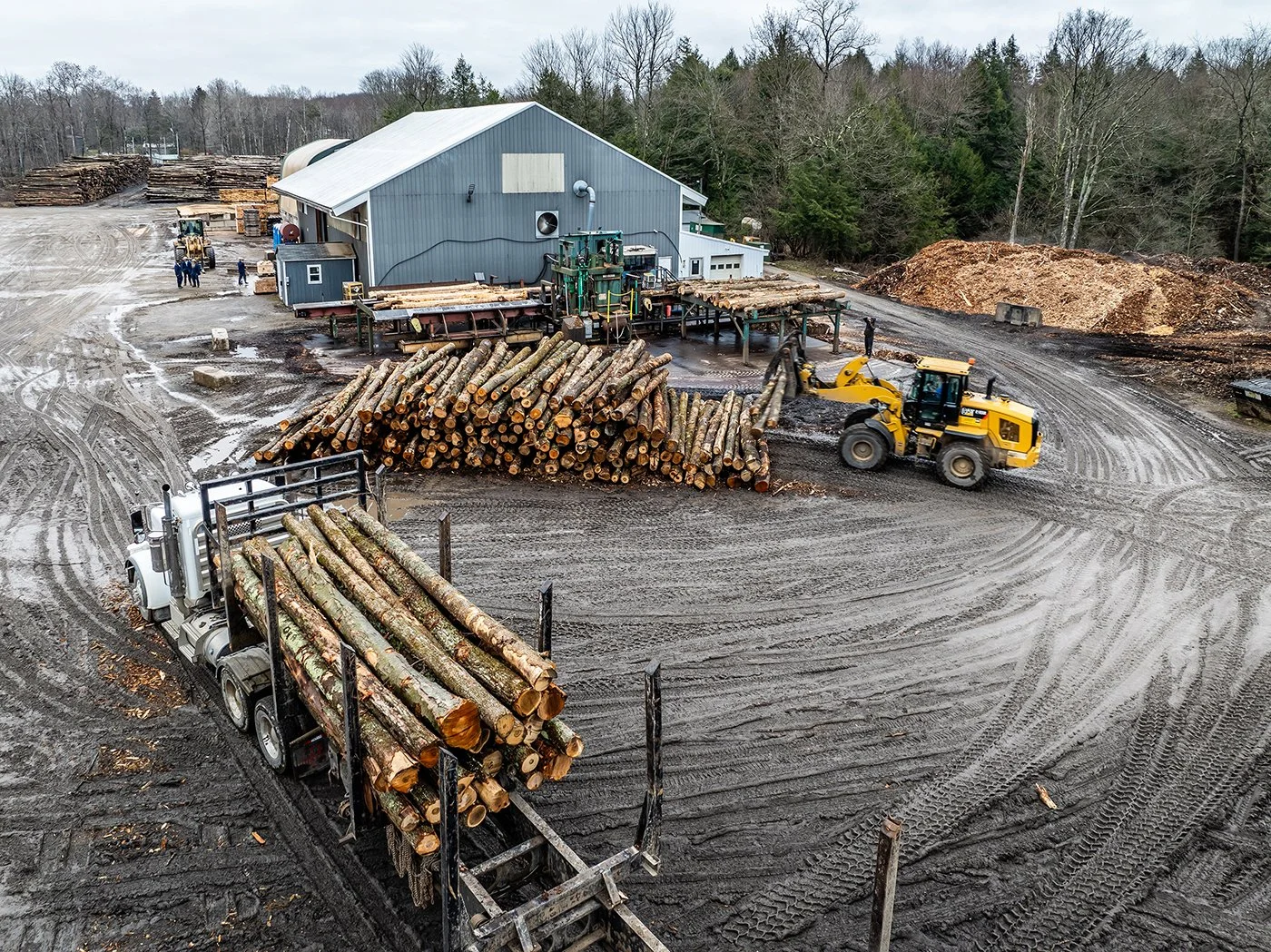 Aerial view of a lumber yard with a stack of logs, a truck loaded with cut limber and loader moving logs