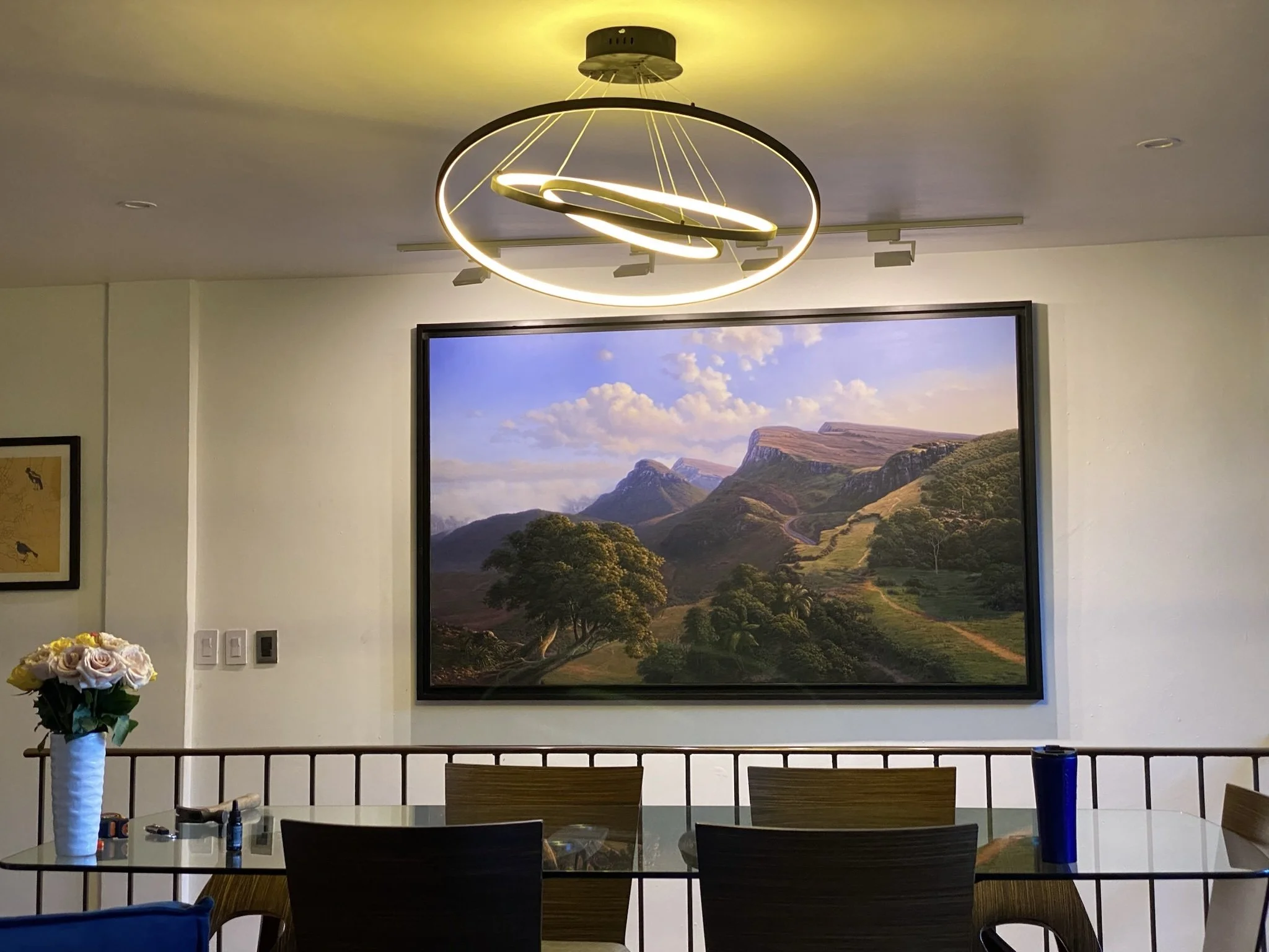 Sala de comedor con cuadro grande de paisaje montañoso en la pared, mesa con sillas, florero con rosas y lámpara moderna en el techo. Casa de las Lomas de Chapultepec. Paisaje contemporáneo cubano. Picci Fine Arts. 