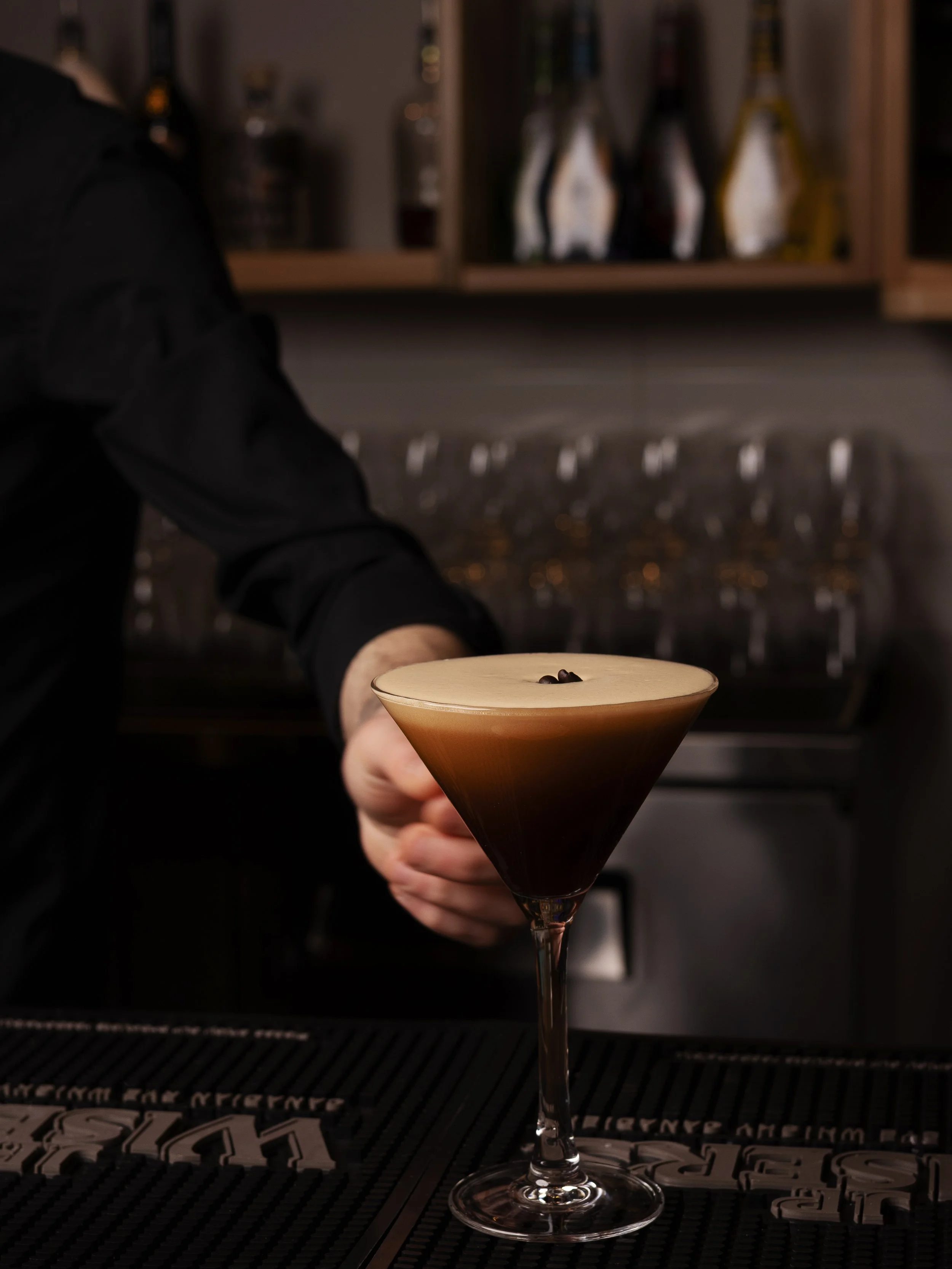 A bartender holding a dark cocktail in an elegant glass, topped with a creamy layer and coffee beans, with a bar background featuring shelves and glasses.