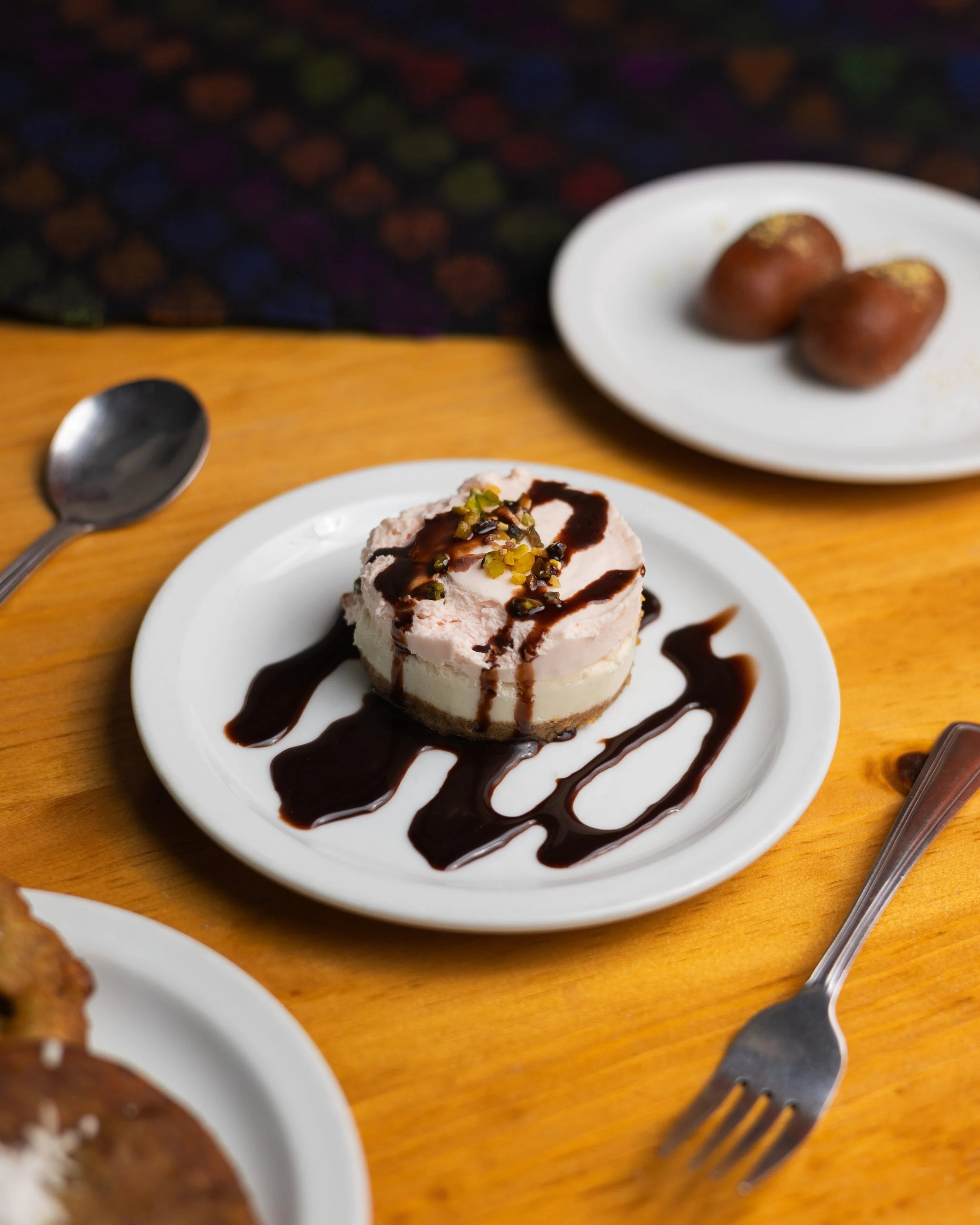 A slice of cheesecake on a white plate, drizzled with chocolate sauce and chopped nuts, on a wooden table.