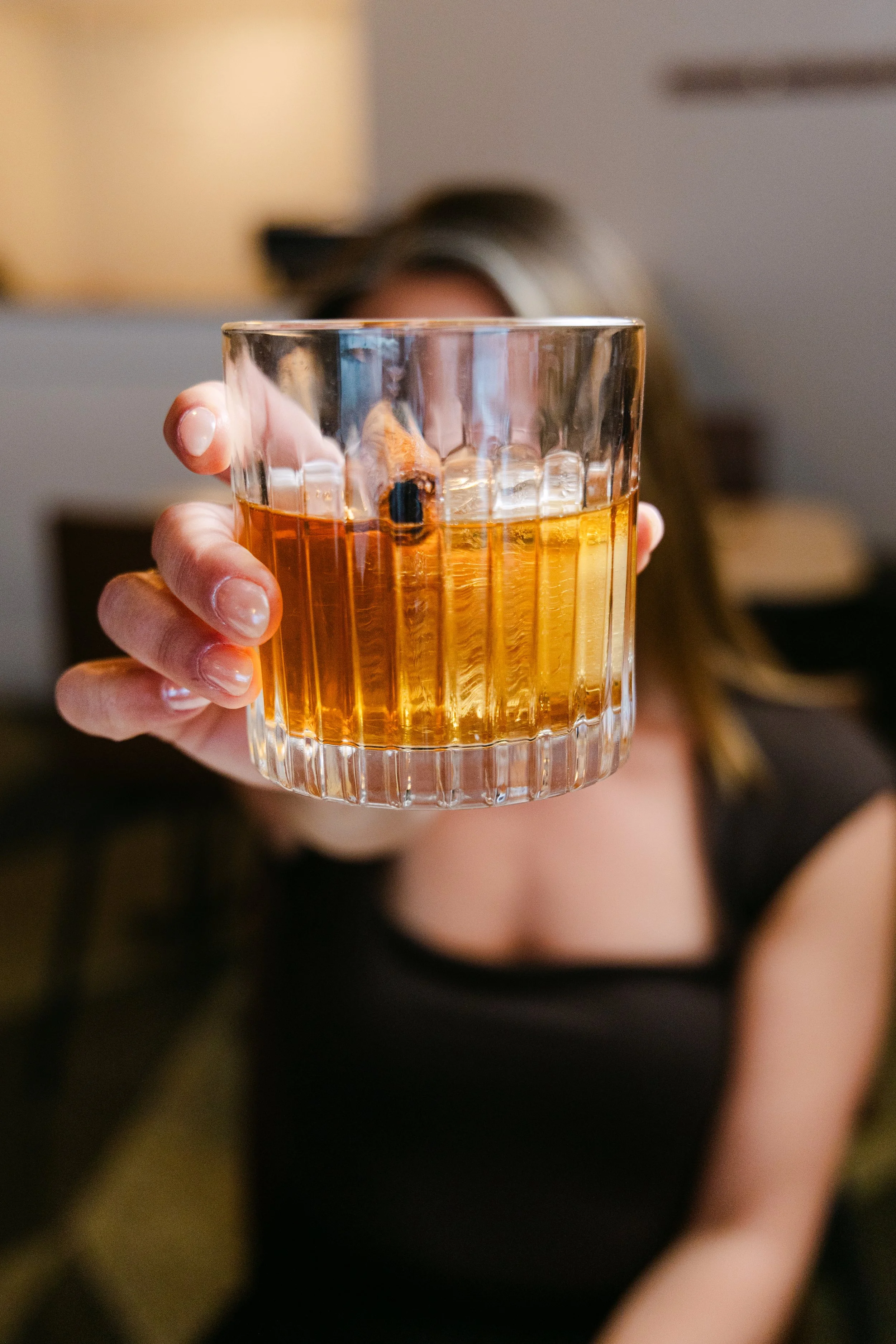 Person holding a glass of amber-colored whiskey in front of their face, with a blurred background.