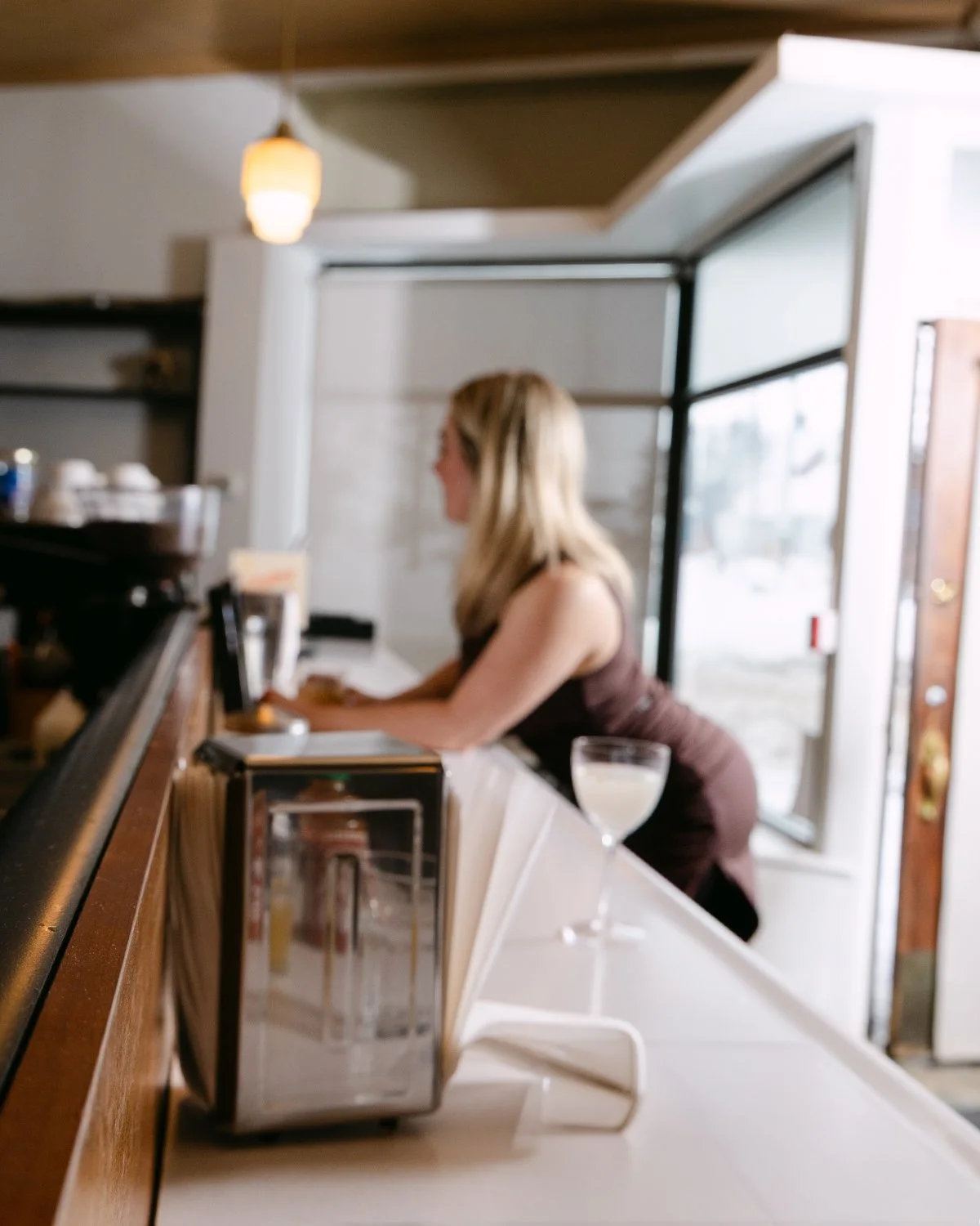 Blurry image of a woman leaning on a bar counter at a restaurant, with a drink in front of her and a window in the background.