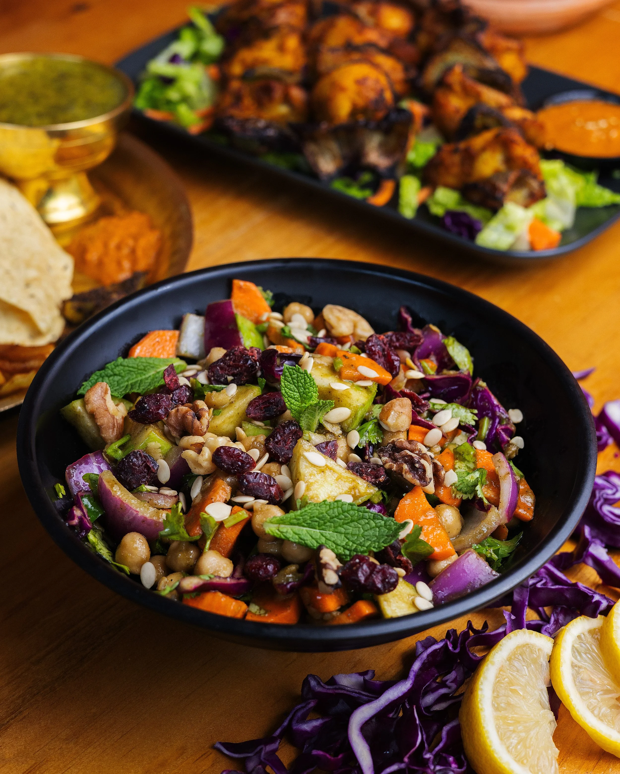 A black bowl filled with a colorful vegetable salad containing chickpeas, carrots, red onions, cucumbers, dried cranberries, sunflower seeds, and fresh mint leaves. In the background, there is a tray of grilled chicken wings, a small bowl of green sa