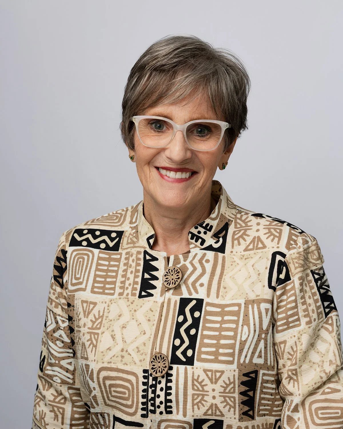 A woman with short gray hair, wearing white glasses, gold earrings, and a patterned beige and black jacket, smiling against a plain light gray background.