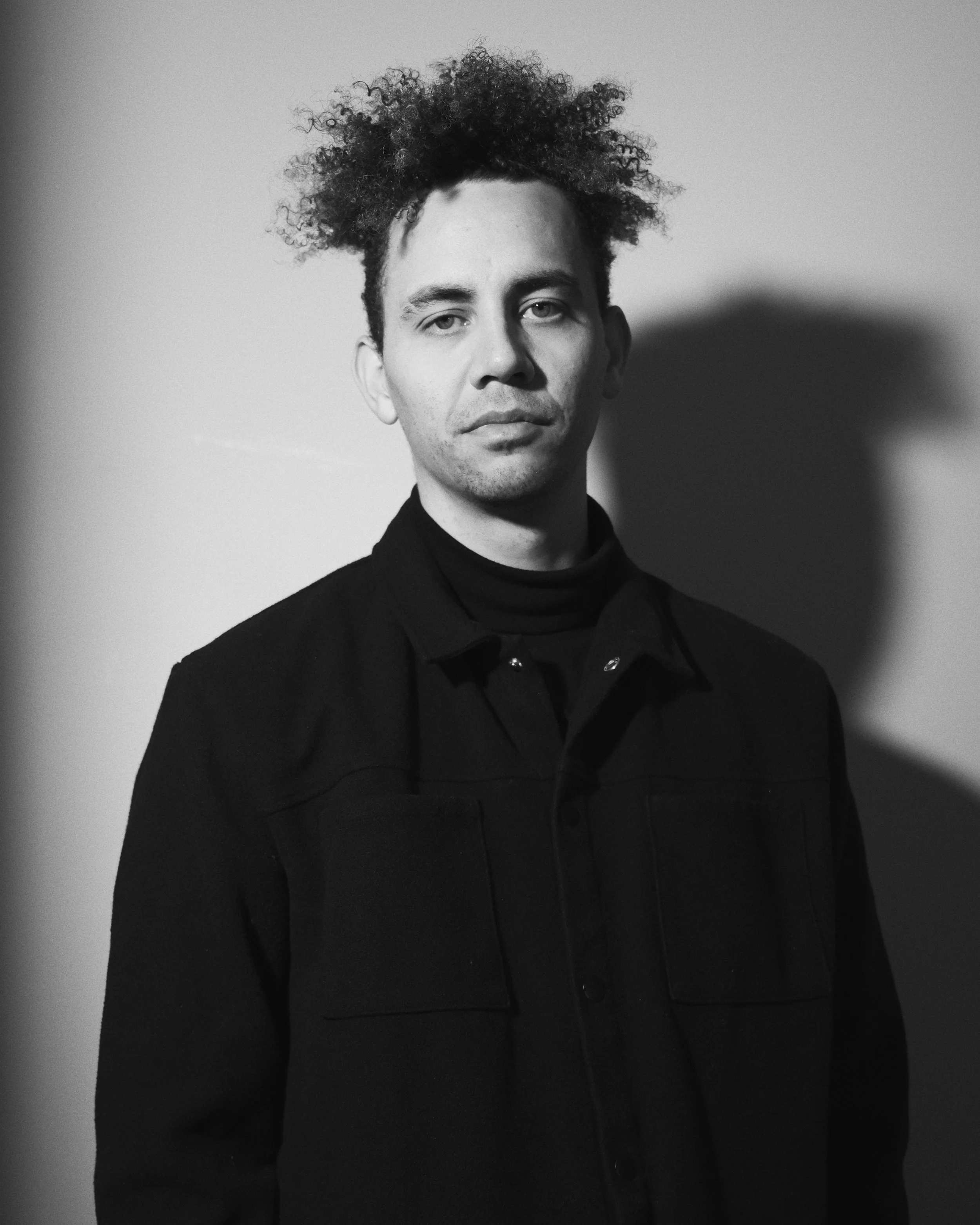 A young man with light skin, short dark hair, and blue eyes, wearing a beige beanie and black shirt, standing indoors near a bright window.