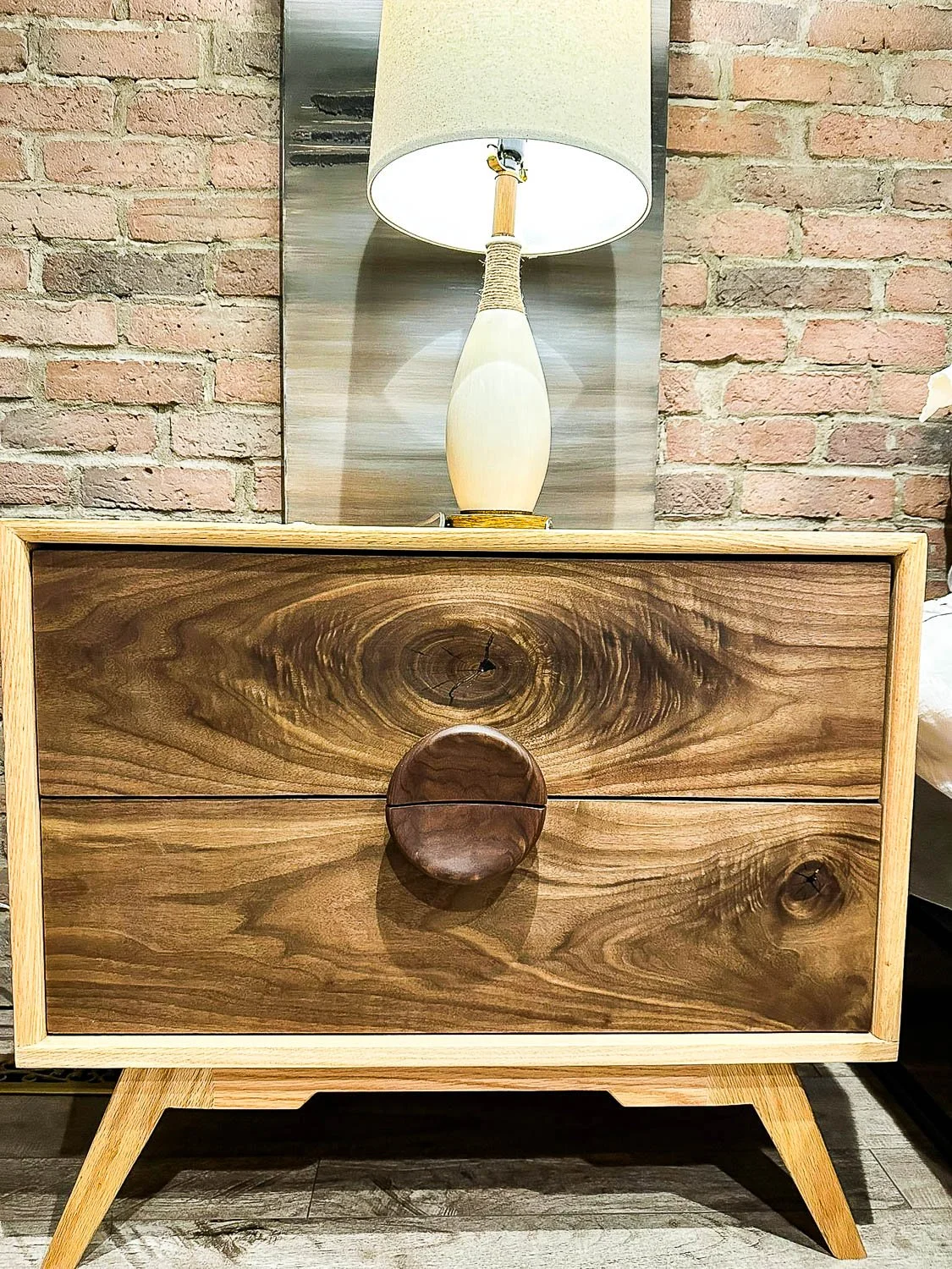 A wooden dresser with two drawers and a large round wooden handle, placed in front of a brick wall. On top of the dresser, there is a lamp with a beige shade and a cream-colored, oval-shaped base.