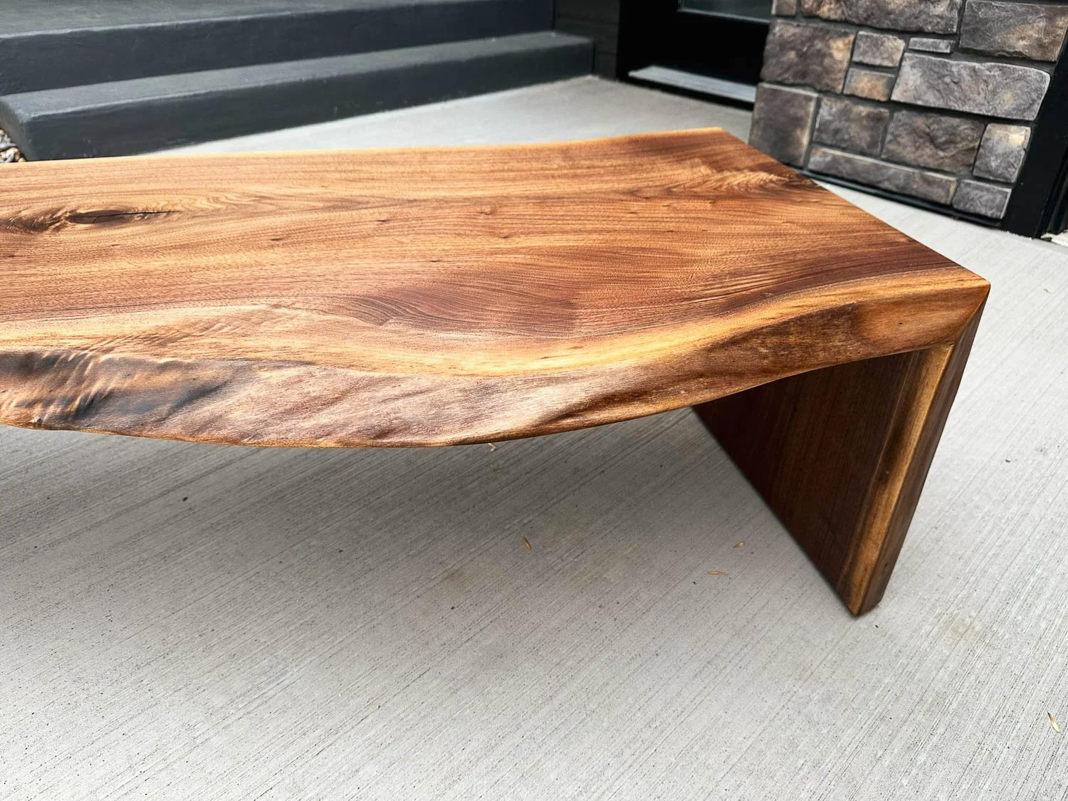 A wooden coffee table with a live edge, placed on a concrete surface outside a house.