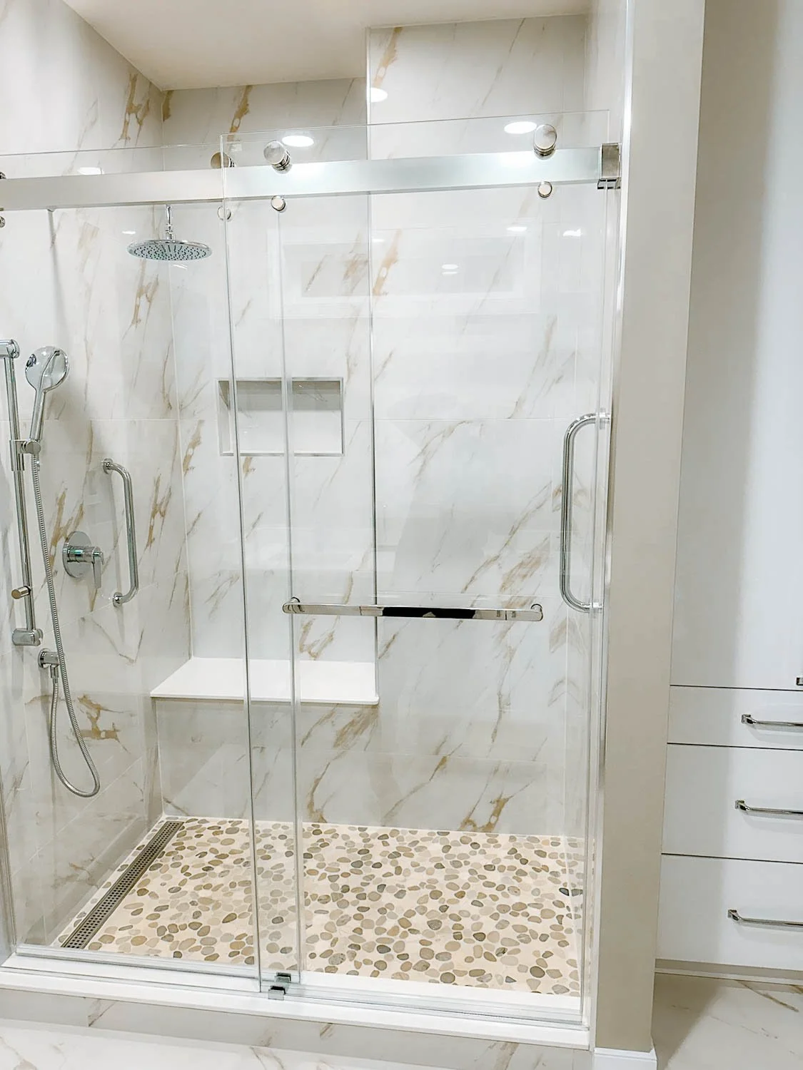 A modern walk-in shower with clear glass sliding doors and marble walls. The shower features a ceiling-mounted rainfall showerhead, a handheld showerhead with a hose, and a built-in shelf. The shower floor is decorated with pebble tiles.