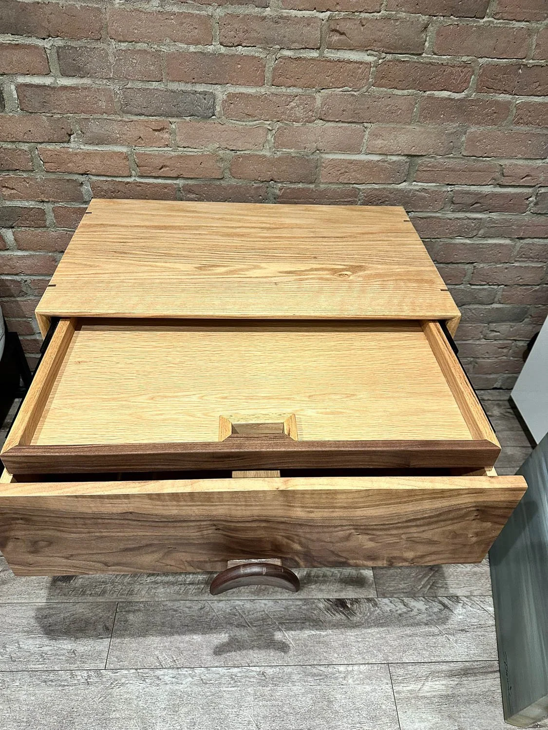 A wooden drawer chest with one drawer pulled out, placed against a brick wall.