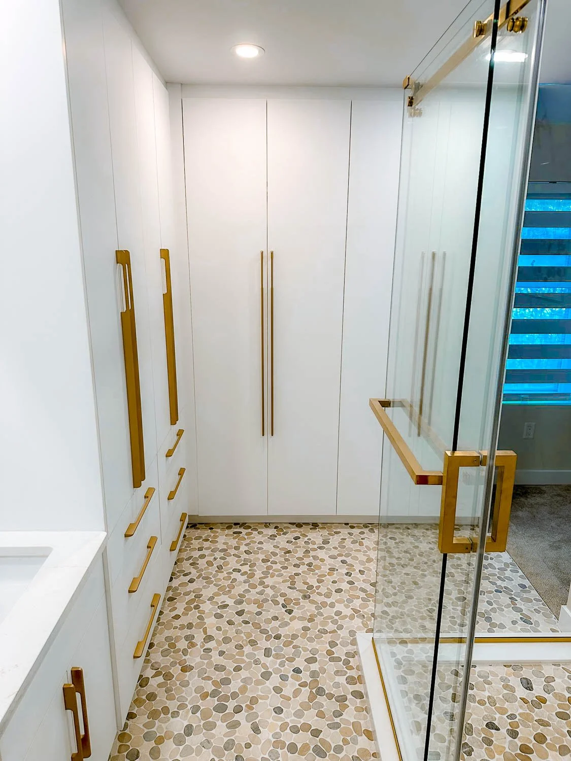Modern bathroom with white cabinets, gold handles, and a glass shower enclosure with gold accents. Pebble tile flooring.