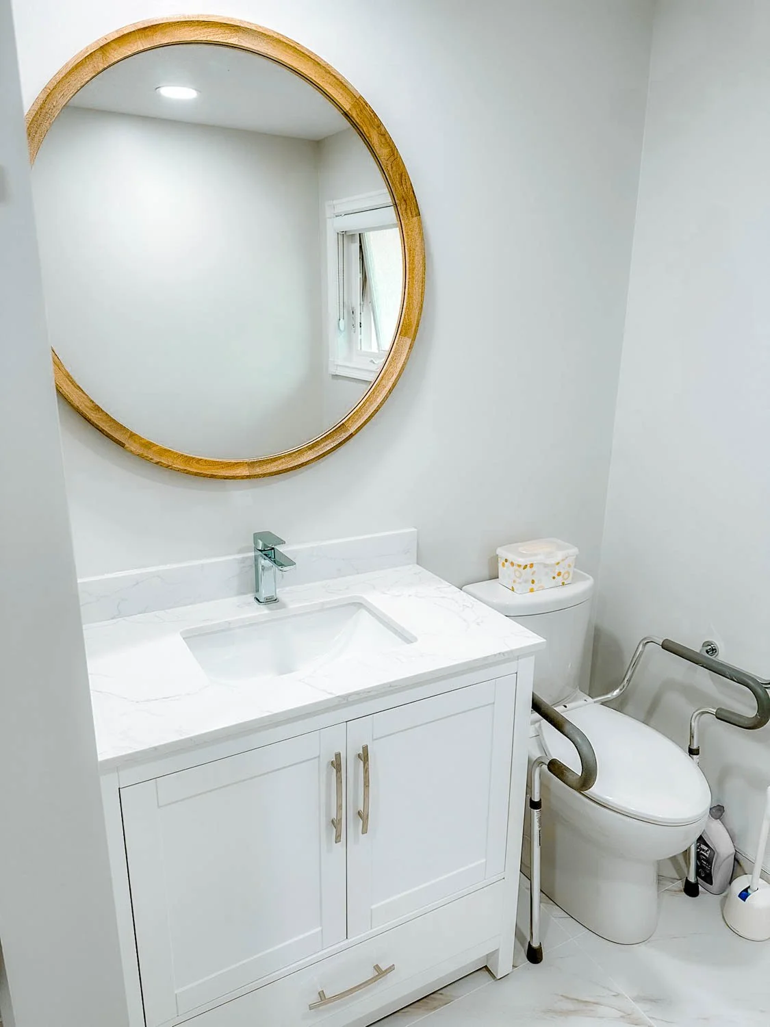 A bathroom with a white vanity with a marble countertop and a rectangular under-mount sink, a round wooden-framed mirror above the vanity, a toilet with safety rails, a small window, and a tissue box on top of the toilet tank.