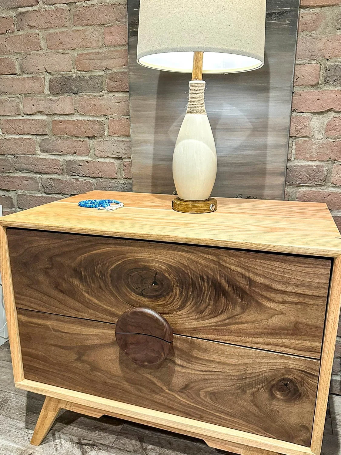 A wooden dresser with a circular wooden handle and a natural wood grain pattern, topped with a ceramic table lamp with a beige shade.