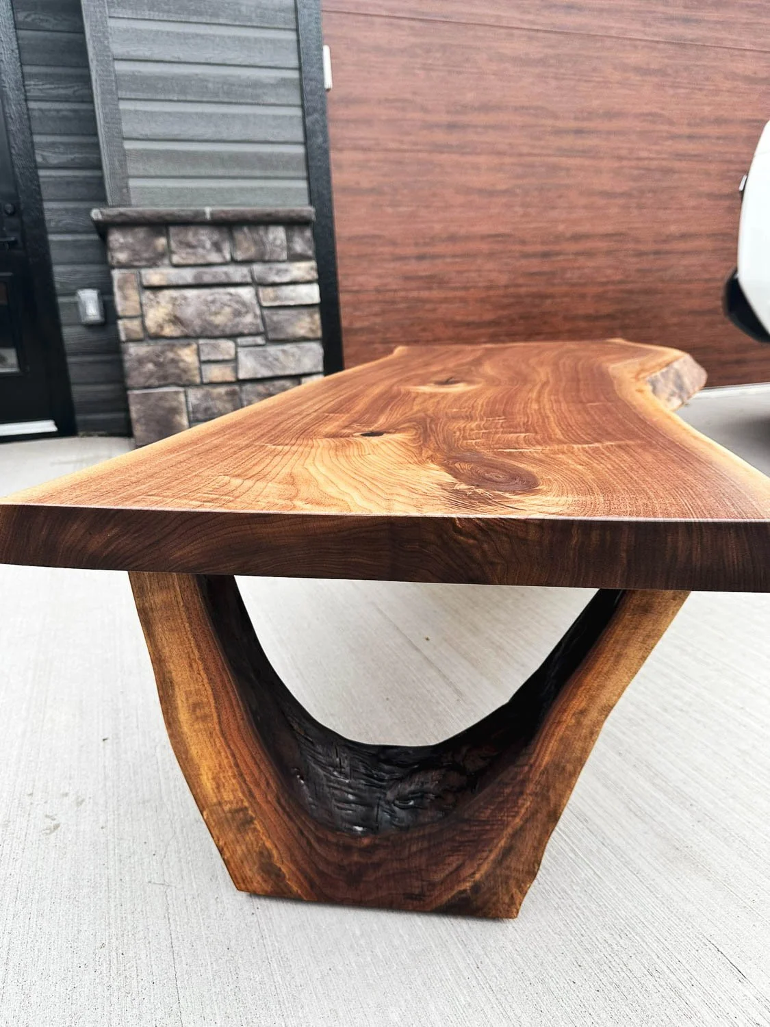 Close-up of a wooden coffee table with a natural edge, showcasing the wood grain and a black center support, set on a concrete surface with a building wall in the background.