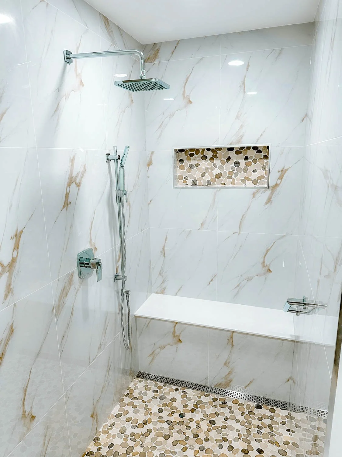 A modern walk-in shower with large white marble-like tiles, a pebble tile accent niche, a bench, and a rainfall showerhead.