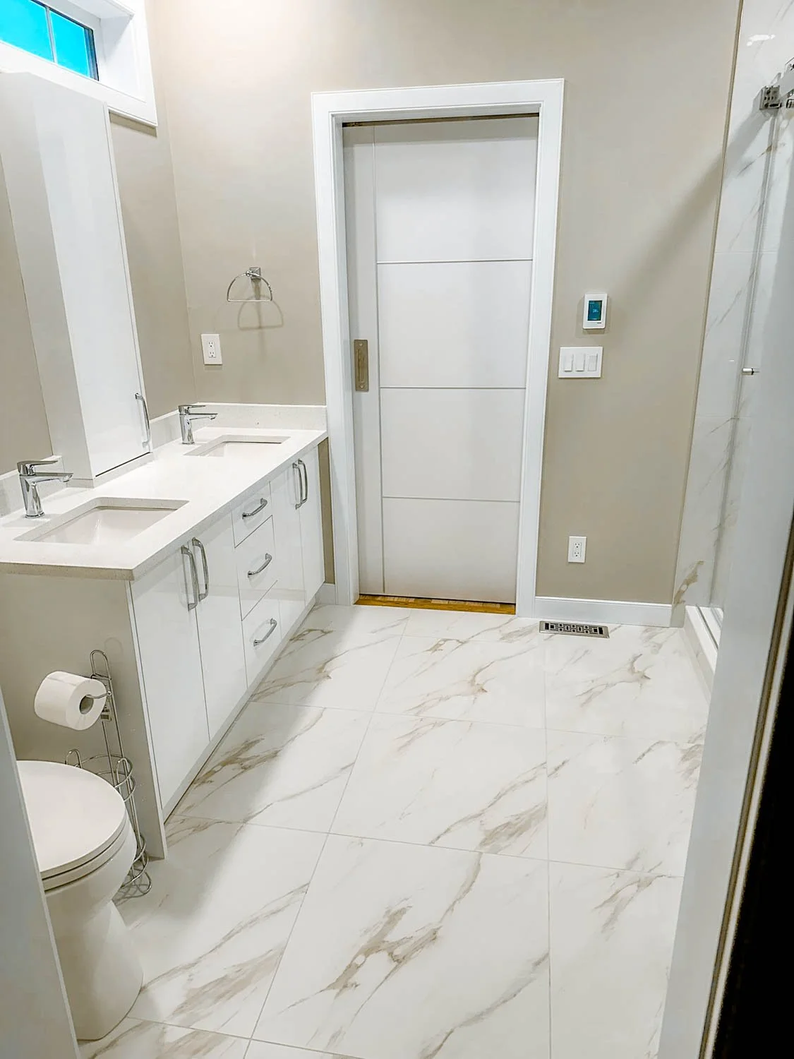 Modern bathroom with double vanity sinks, large marble tiles, a toilet, a sliding door, and a small window.