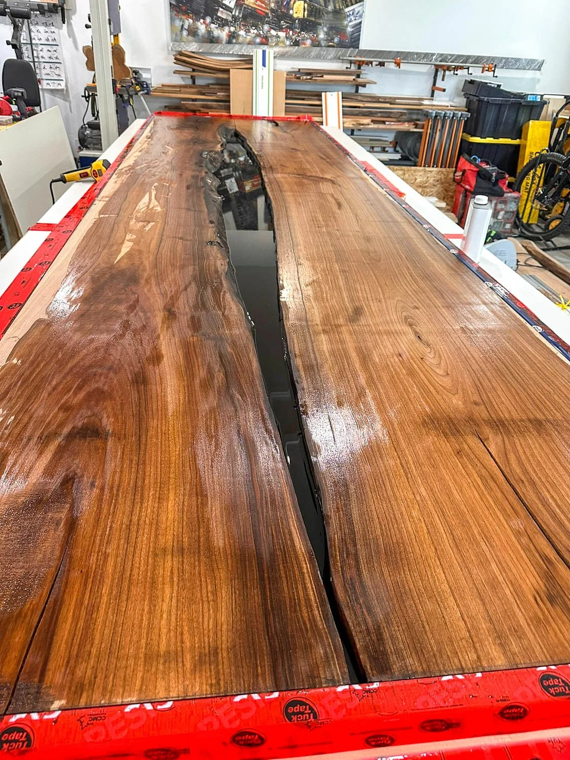 A large, polished wooden table with a dark resin river running through the center, situated in a woodworking workshop surrounded by tools, wood pieces, and storage bins.