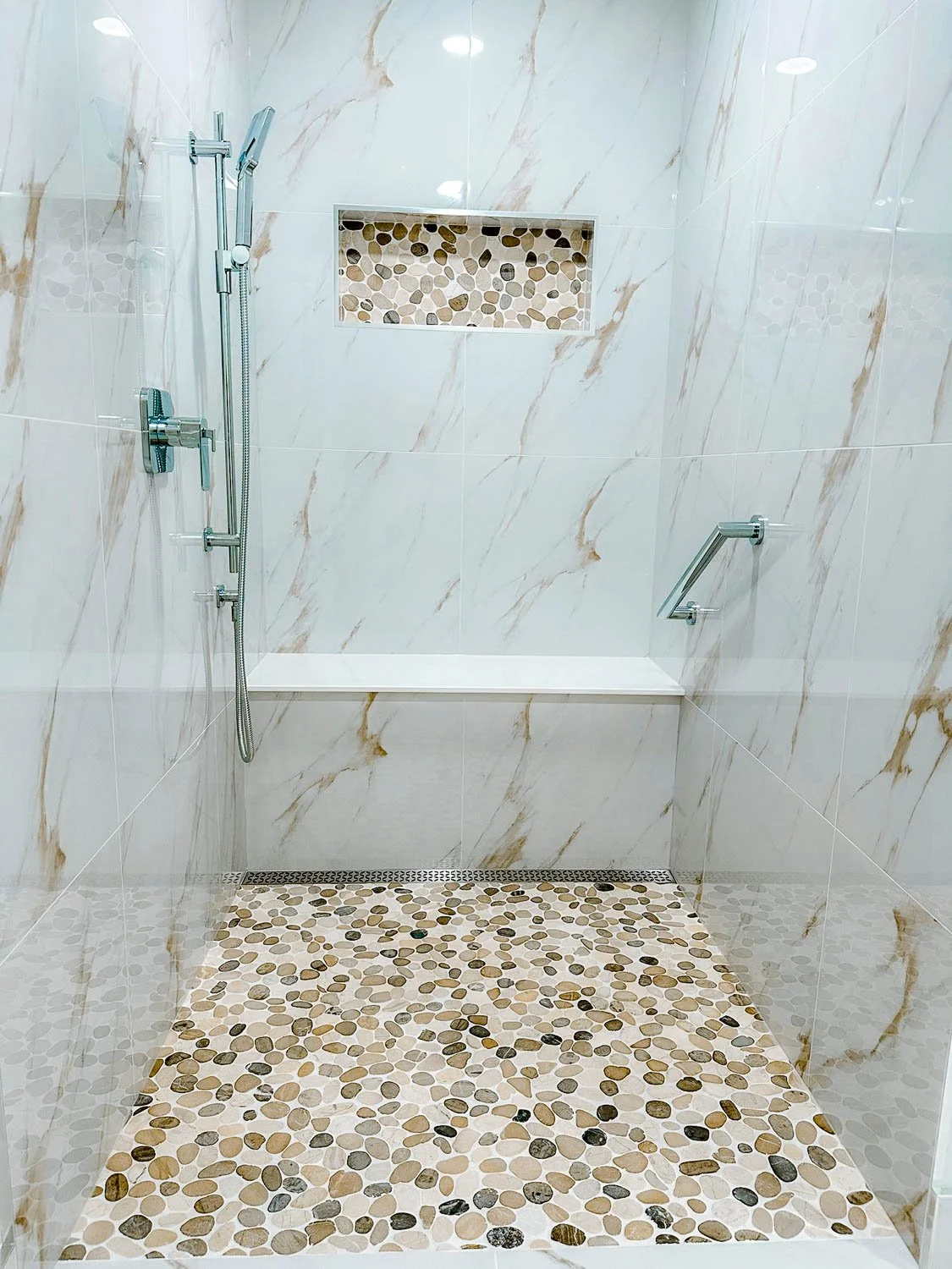 A walk-in shower with a mosaic pebble floor, white marble wall tiles with gold veining, a built-in pebble niche, a chrome hand-held shower, and a metal grab bar.