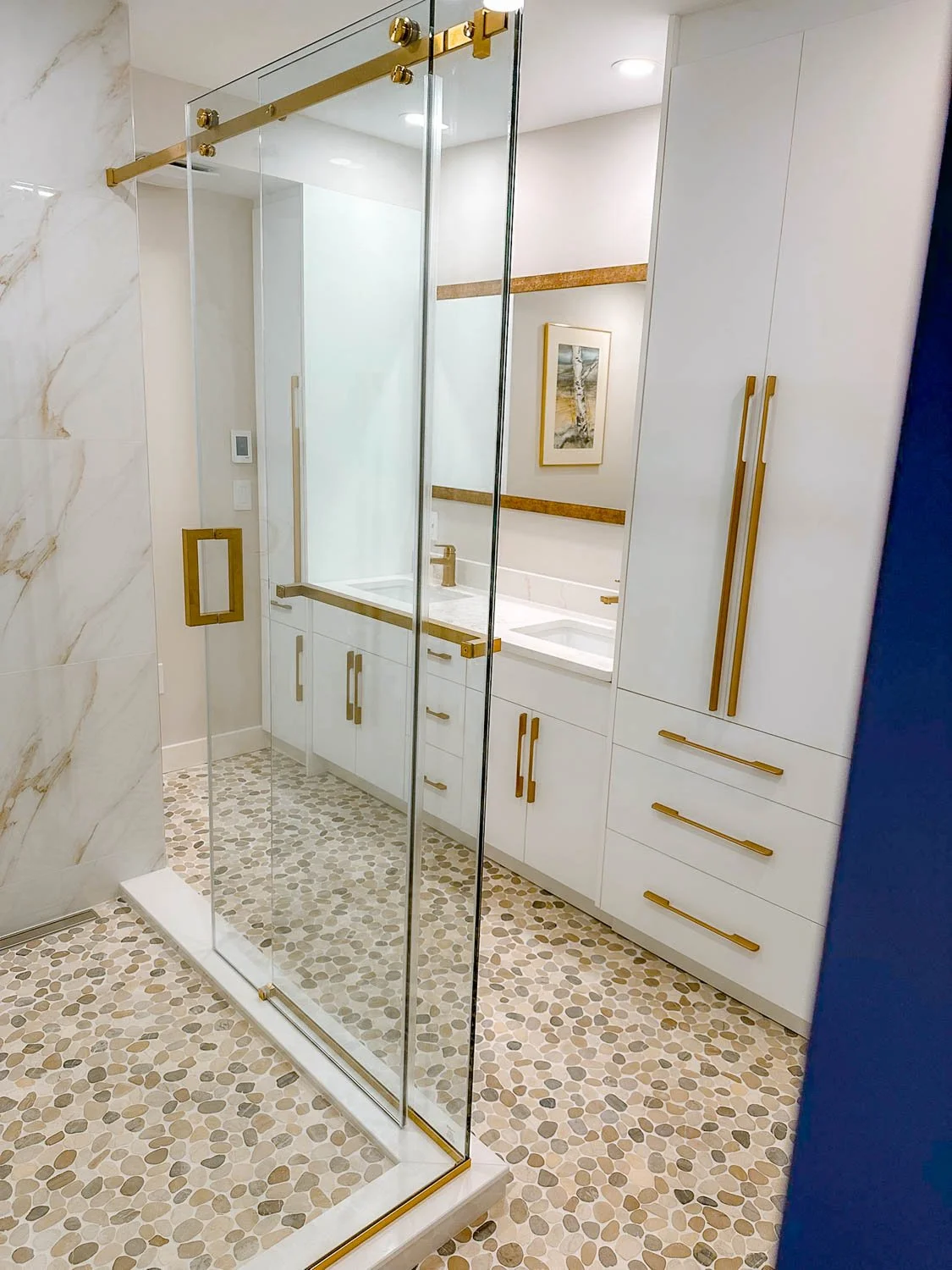 Modern bathroom with glass shower enclosure, white cabinets with gold handles, and a marble countertop. The shower floor has pebble tiles, and there is a framed artwork on the wall.