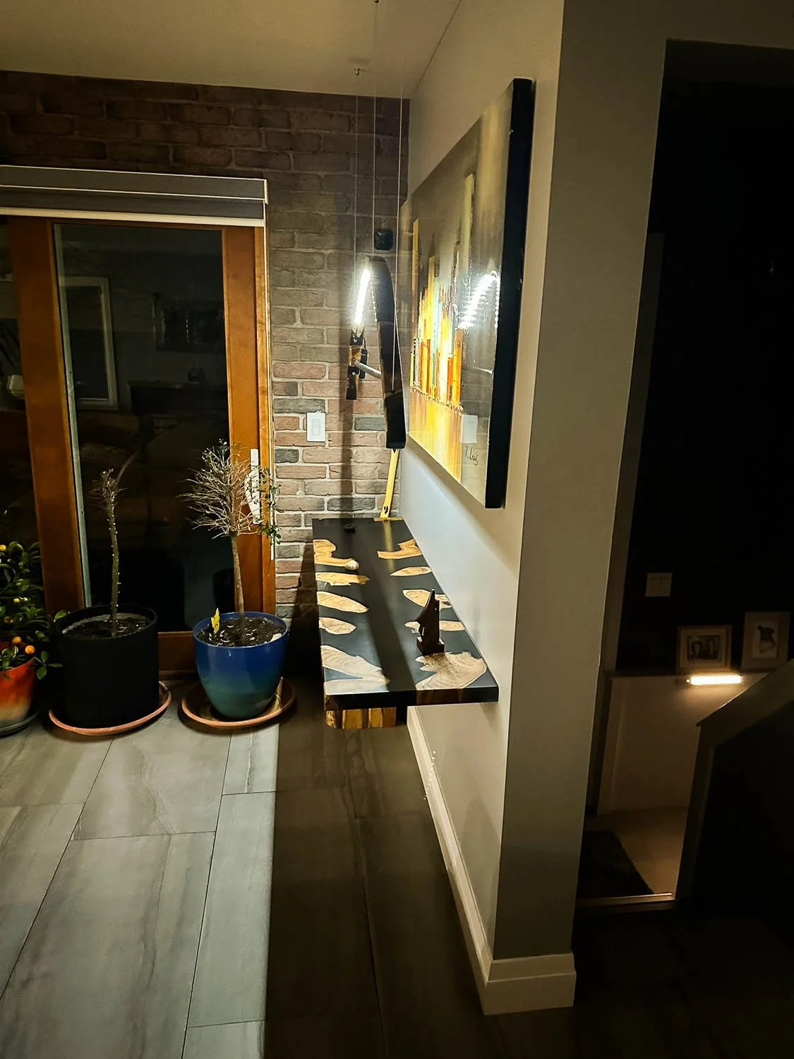 Indoor scene with a decorated console table against a cream wall, featuring small sculptures, with framed artwork, potted plants, and a brick wall background.