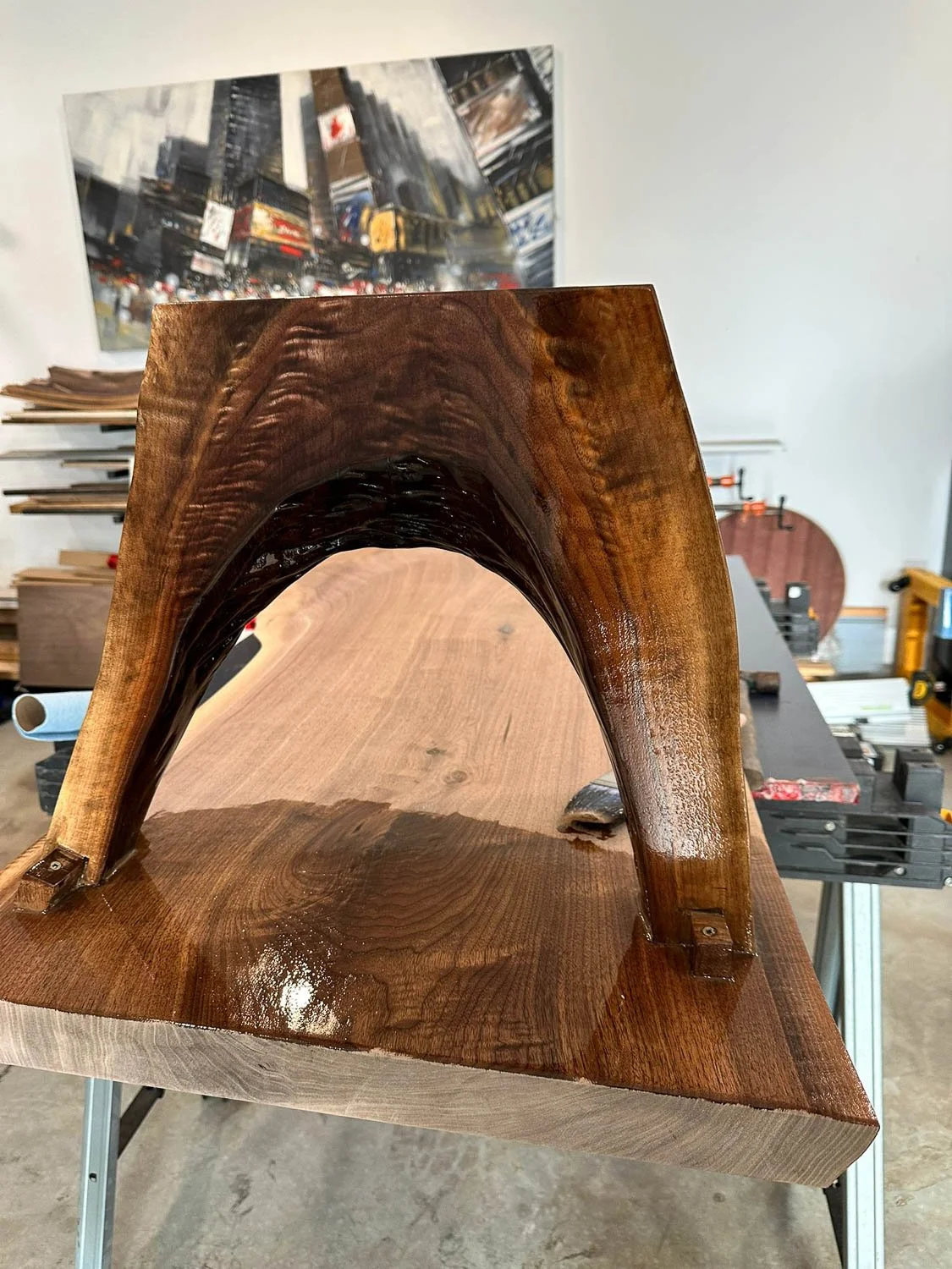 A wooden sculpture or furniture piece featuring a smooth, polished arch shape with a glossy finish, placed on a worktable in a workshop setting.