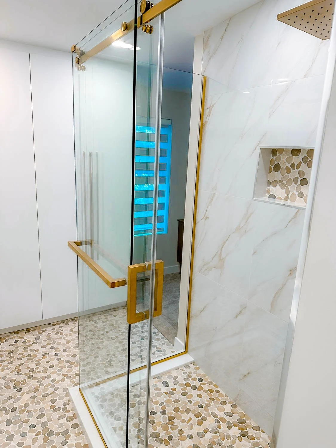 A modern glass shower with a gold handle and trim in a bathroom with pebble floor tiles and a built-in wall niche with small stones.