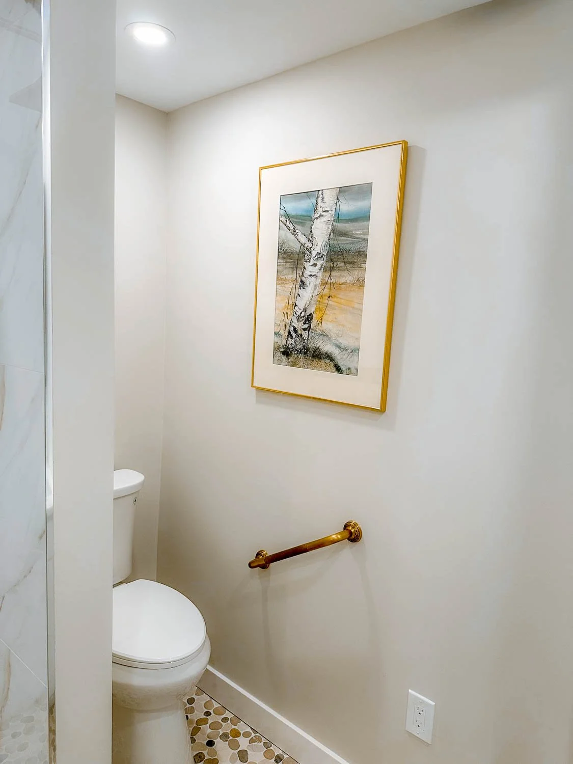 A small bathroom with a white toilet, a framed landscape painting, a wooden grab bar, and a pebble tile floor.
