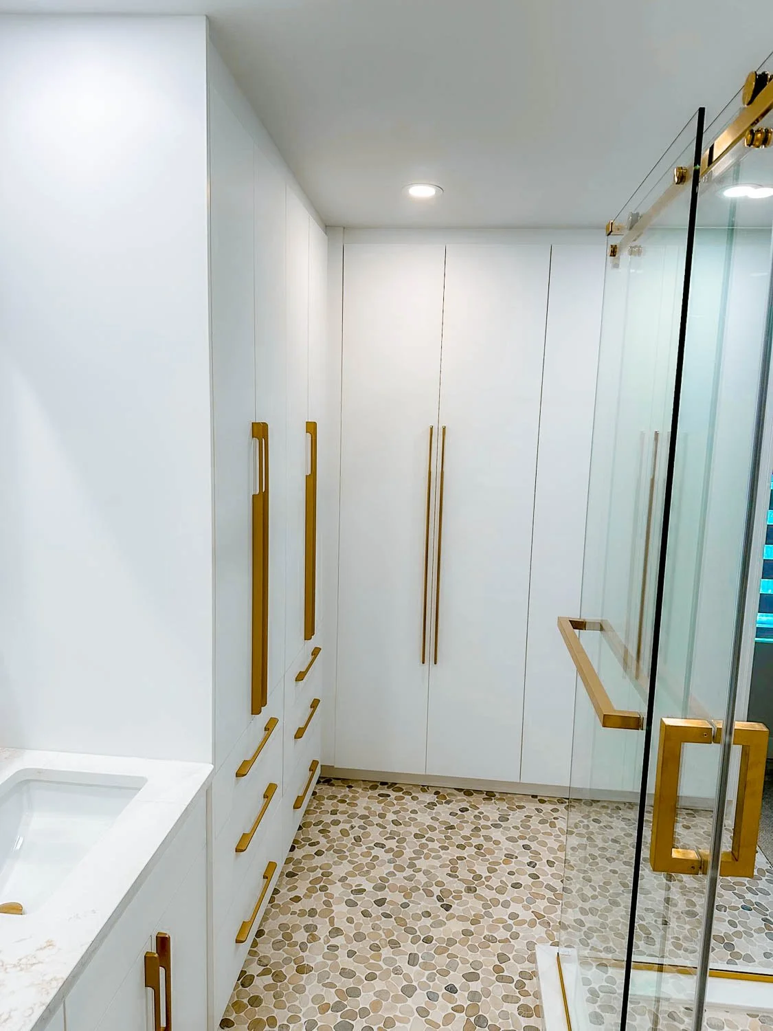 A modern bathroom with white cabinets, gold handles, pebble stone floor, a marble countertop with a sink, and a glass shower enclosure with gold accents.