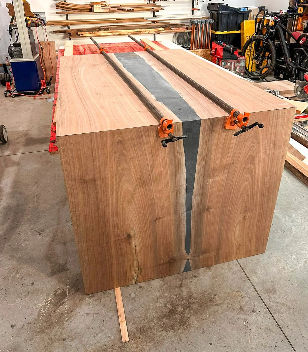 A large wooden slab being glued and clamped in a woodworking shop, with clamps securing black epoxy between the boards.