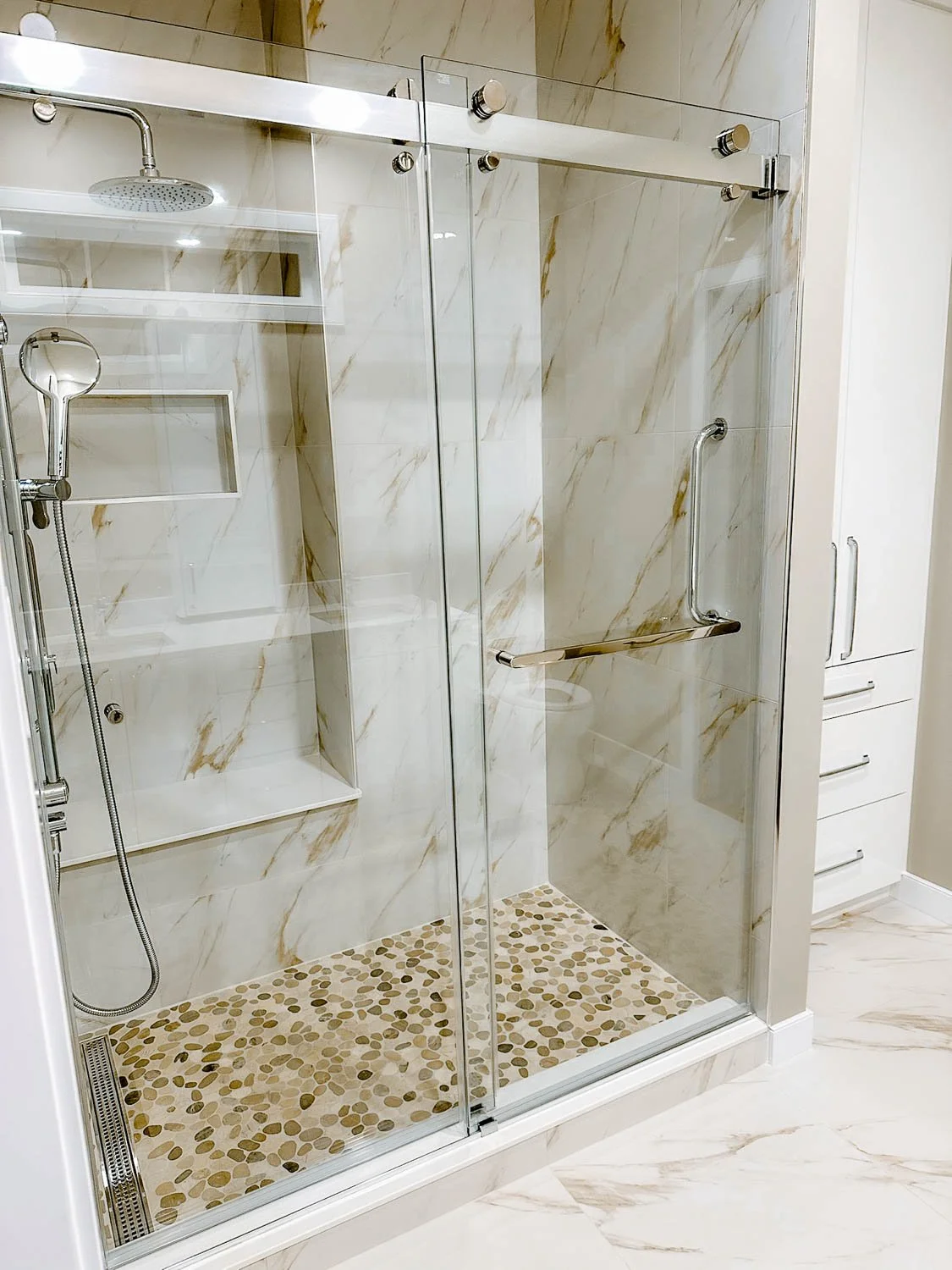 A glass-enclosed shower with marble walls and pebble tile flooring, with a built-in shelf and a handheld showerhead.
