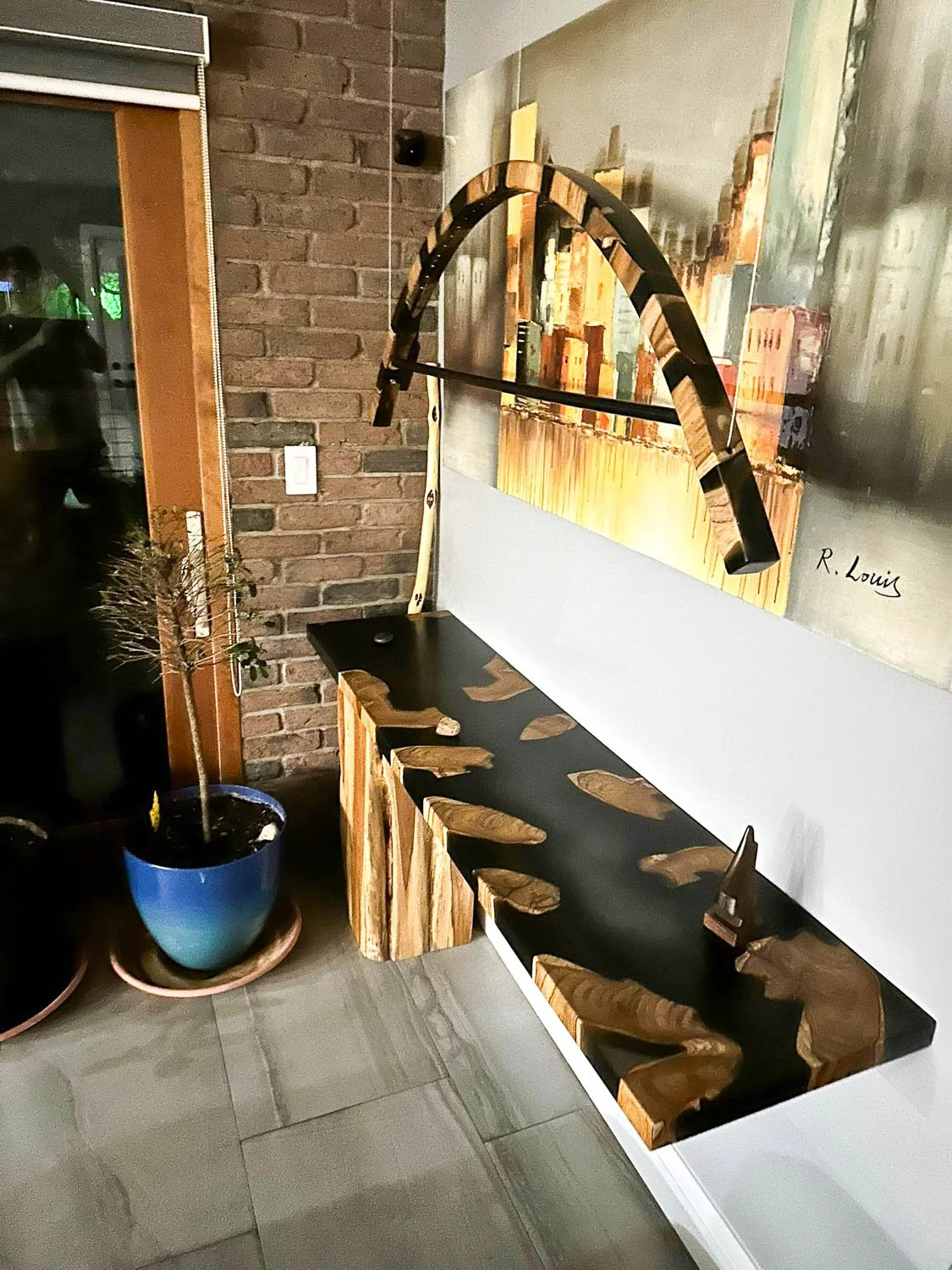 A black wooden desk with irregularly shaped wooden inlays, positioned against a white wall with a large abstract painting. Over the desk, a wooden sculpture hangs from a metal rod, resembling a boomerang or arch. To the left, there's a tall potted pl