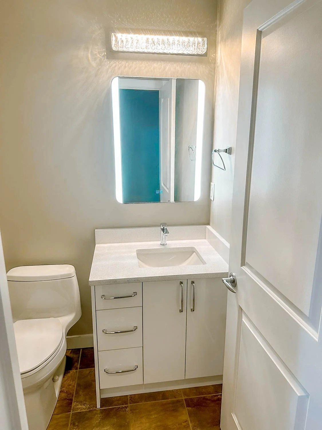 Small bathroom with a white toilet, a white vanity with a sink, a rectangular mirror with LED lighting, and a door.