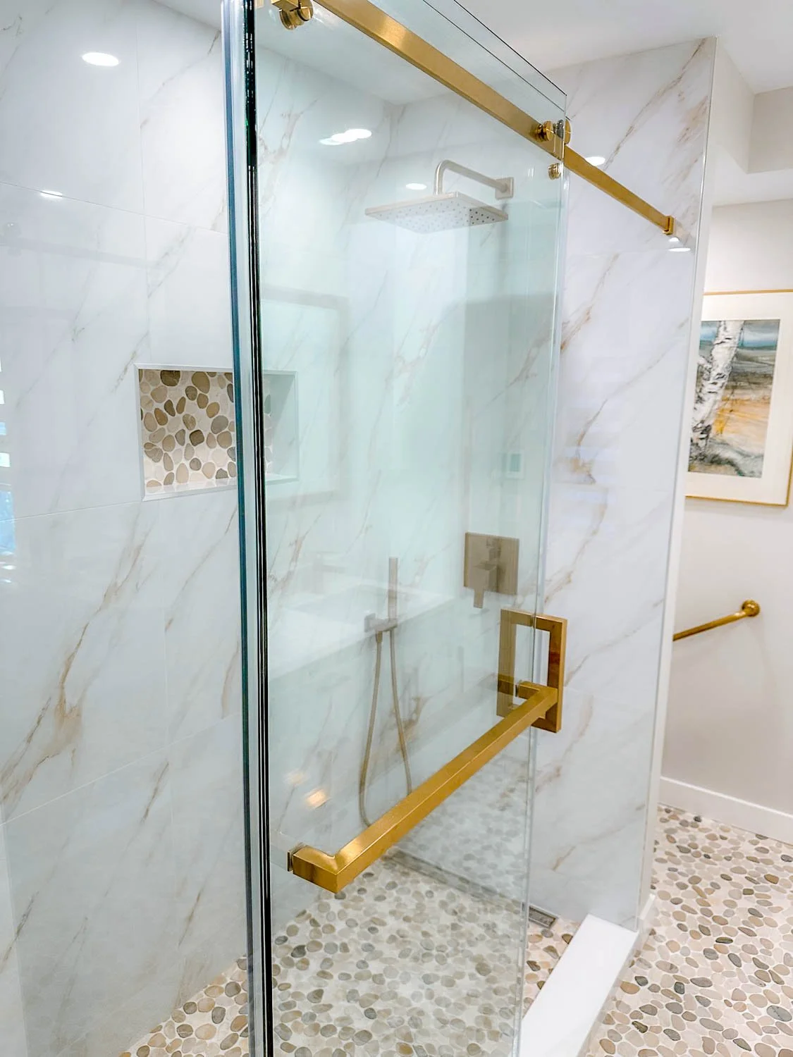 Shower with glass door and gold hardware, marble walls, pebble tile floor, built-in shower niche with pebble pattern, rain showerhead, and handheld shower.