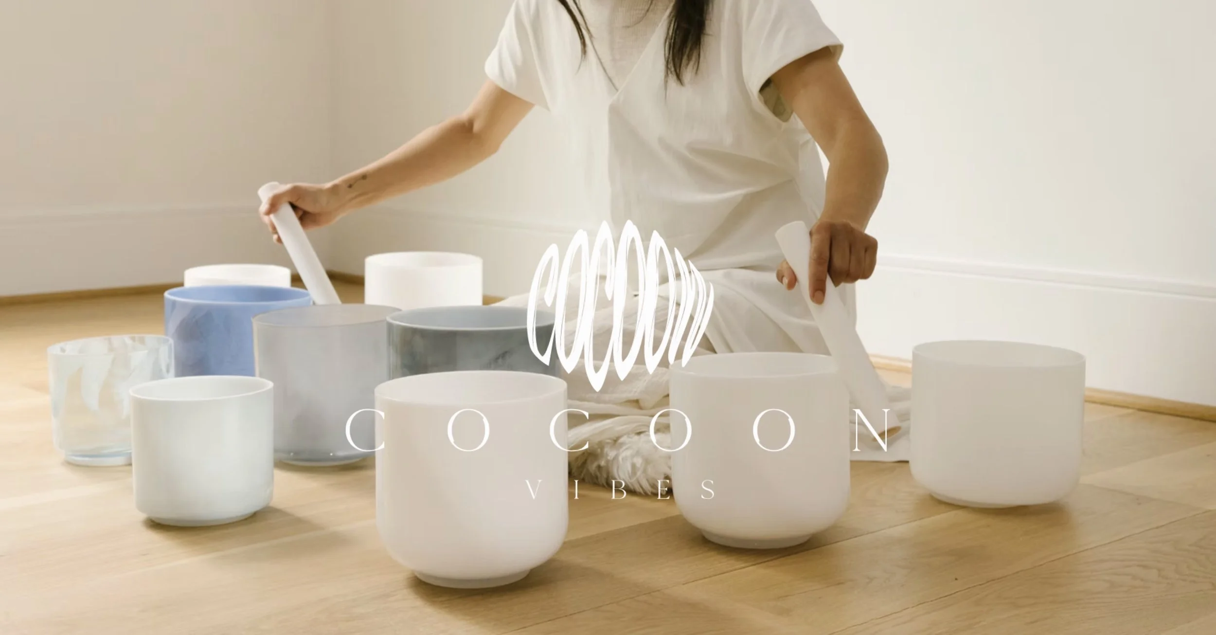 A person sitting on a wooden floor surrounded by various ceramic bowls and cups in different shades of white, gray, and blue, with a white graphic design and text overlay reading "COCOON VIBES."