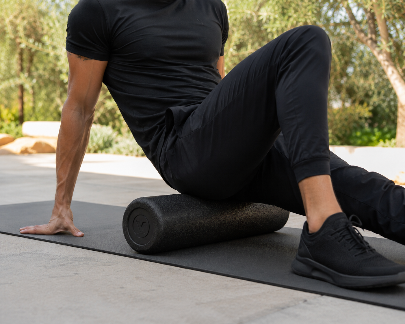 Person using a foam roller on their thigh while seated on a yoga mat outdoors.