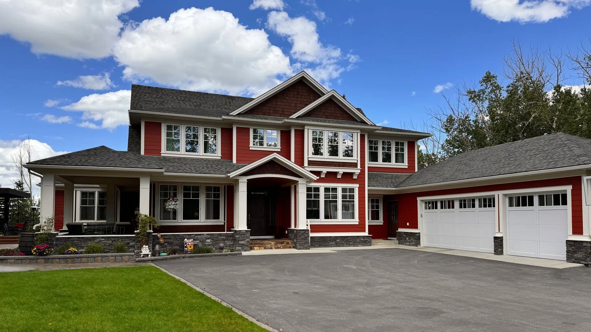 A two-story house with red siding, white trim, and a gray shingle roof, featuring a three-car garage, front porch with stone accents, and landscaping with flowers and greenery, under a partly cloudy sky. Exterior painting of siding, trim, and garage.
