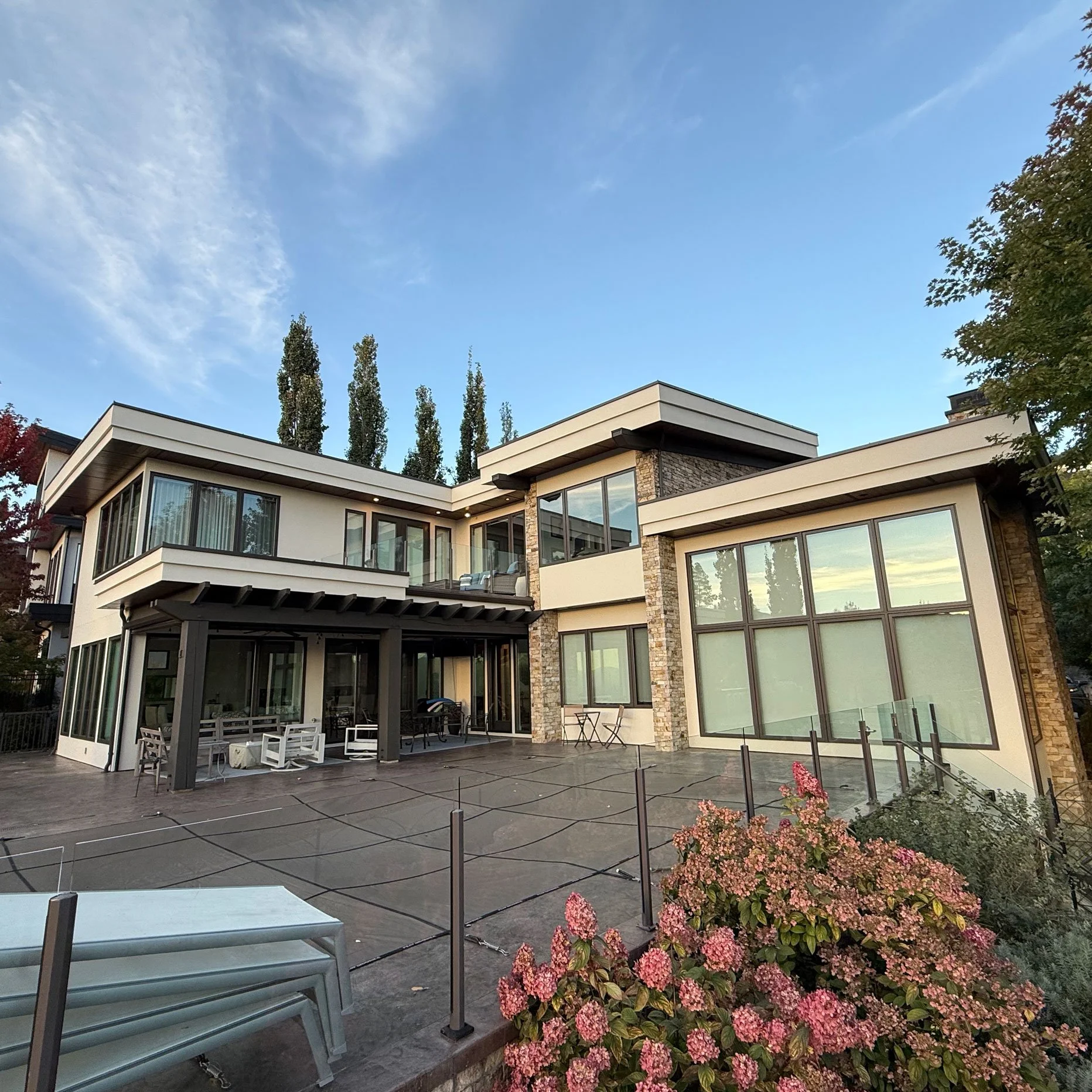 Modern multi-story house with large glass windows, balconies, and a backyard patio with outdoor furniture, surrounded by flowering plants and tall trees under a partly cloudy sky.