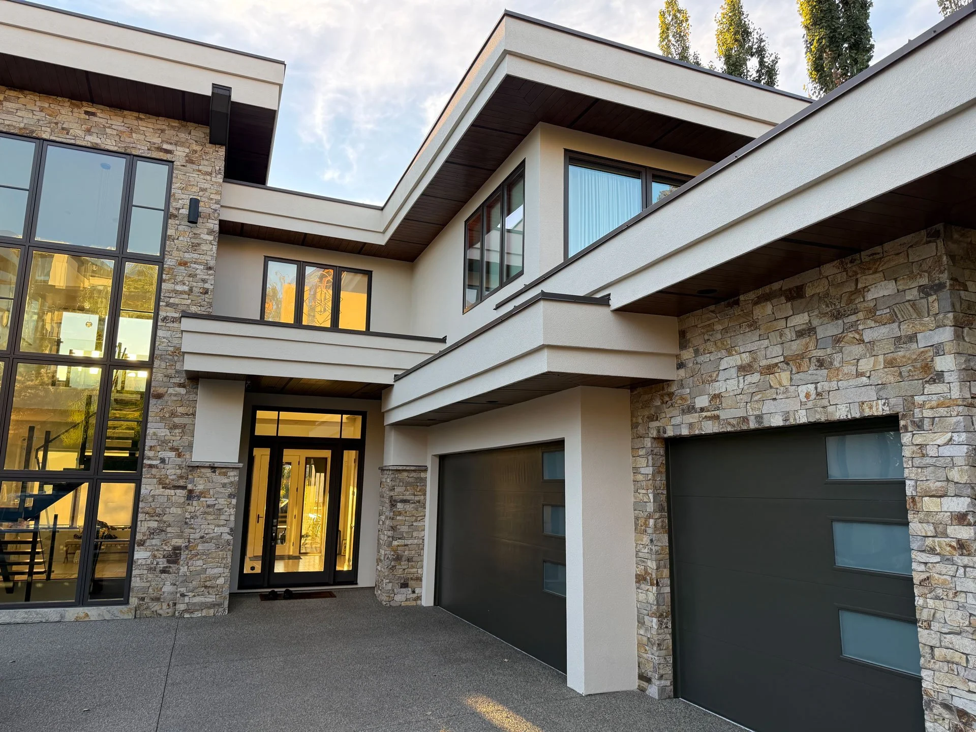 Modern multi-story house with stone and stucco exterior, large windows, and a garage, surrounded by trees.