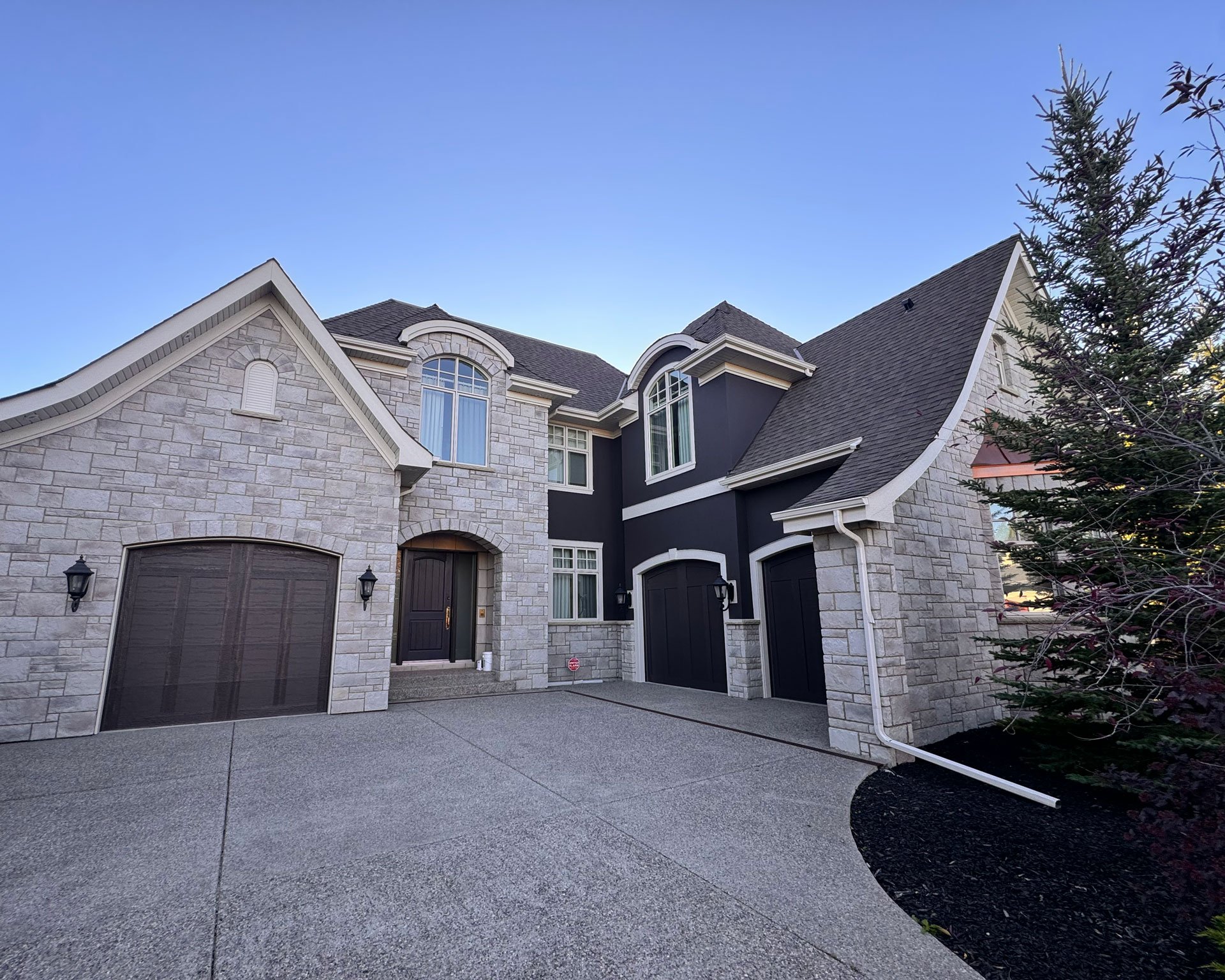 Front view of a modern house with stone and dark siding, two garages, large windows, and a small tree on the right. The driveway is paved and clean.