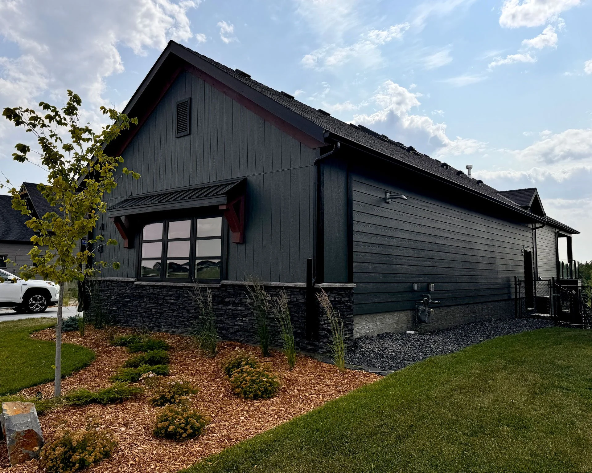 A modern dark gray house with a sloped roof, large front window, and a landscaped front yard with young trees and bushes, under a partly cloudy sky.