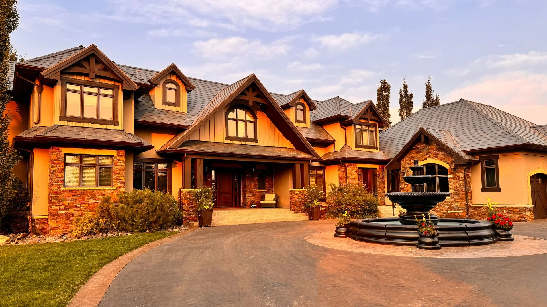 A large, elegant house with a stone and stucco exterior, multiple gabled roofs, and a front porch featuring a fountain and landscaped garden at sunset.