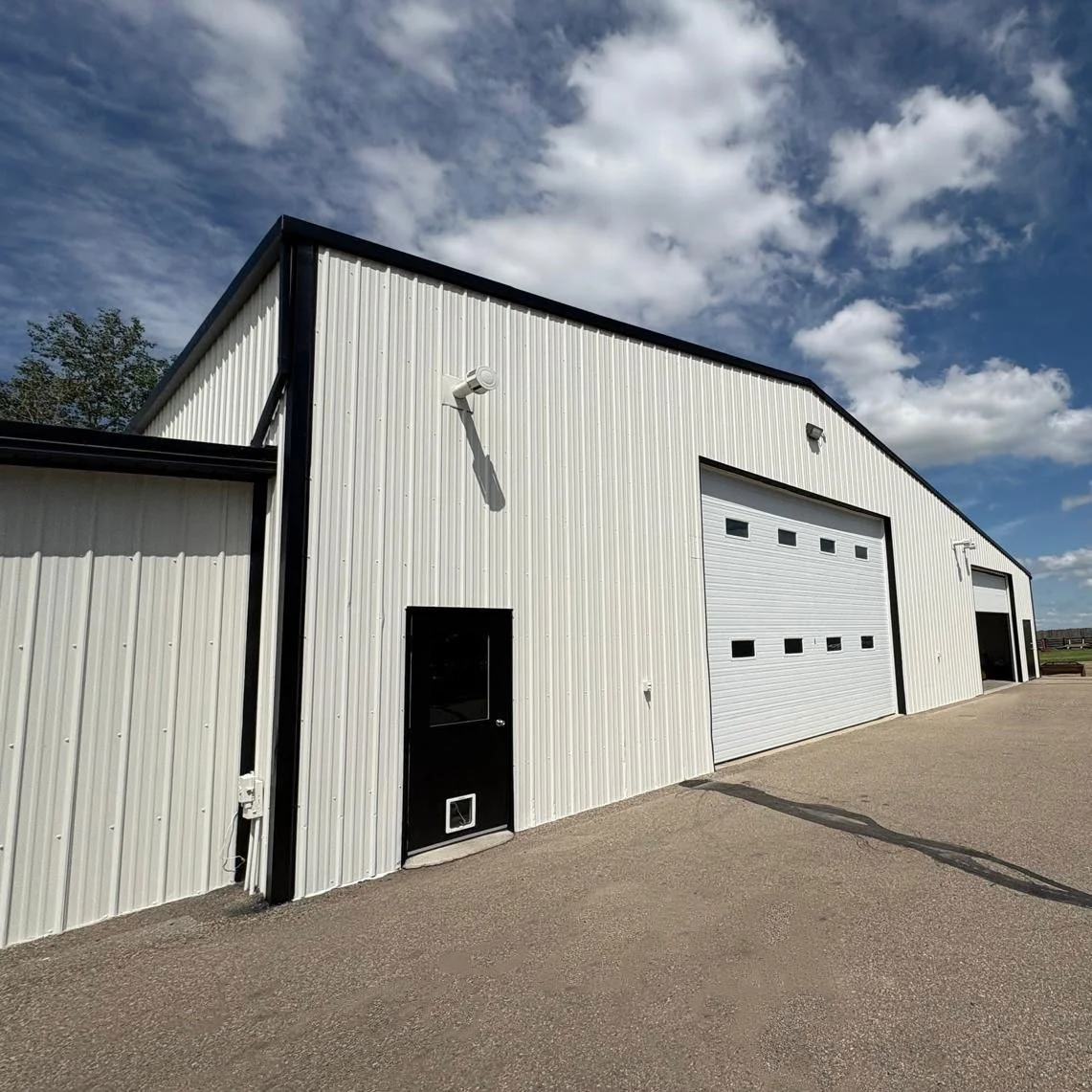 A commercial building freshly painted by JC Paint, white metal siding painted, two large garage doors painted, a small black door, and a security camera under a blue sky with scattered clouds.
