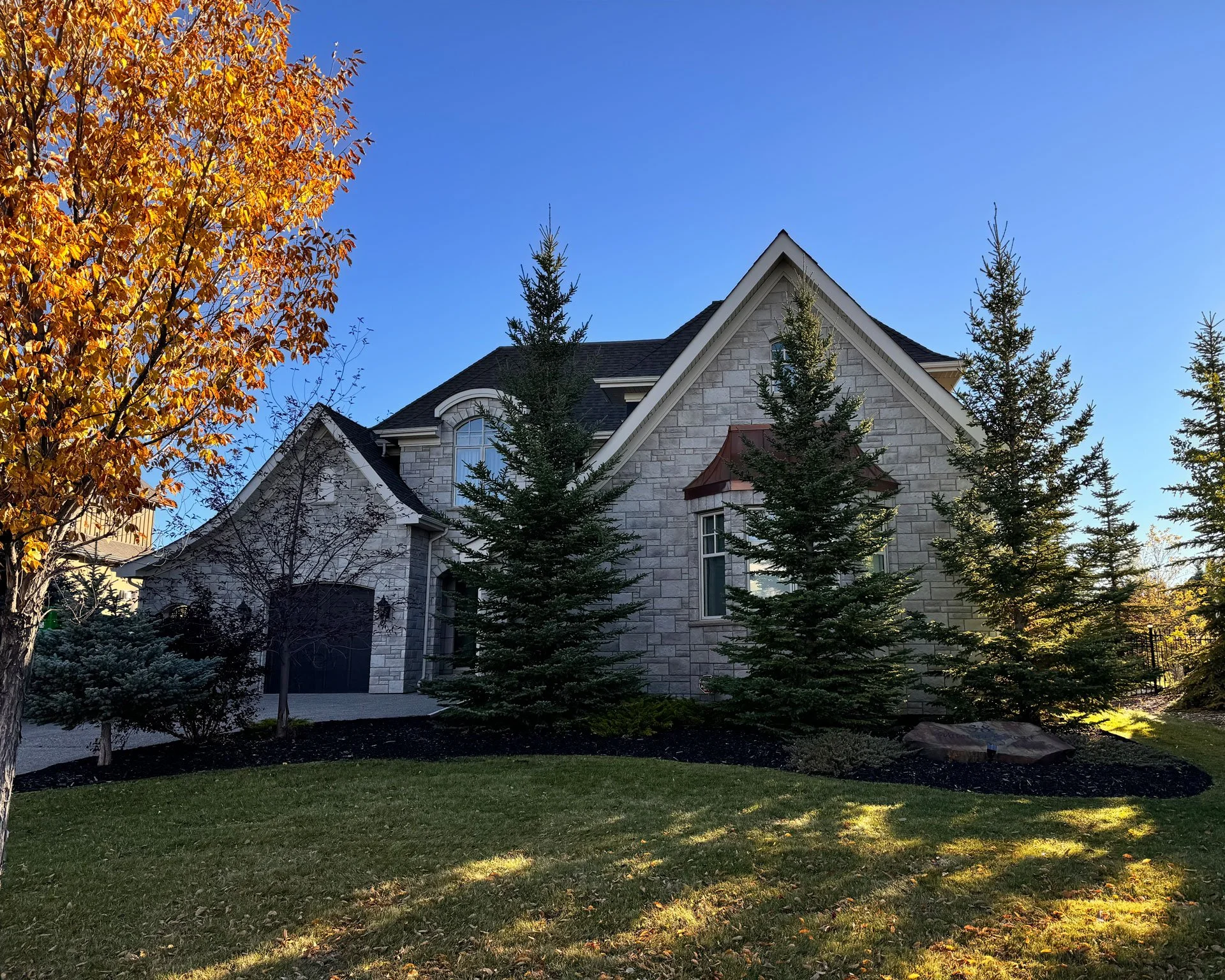 A large stone house with tall peaked roofs, surrounded by green trees and a grassy yard, set against a bright blue sky.