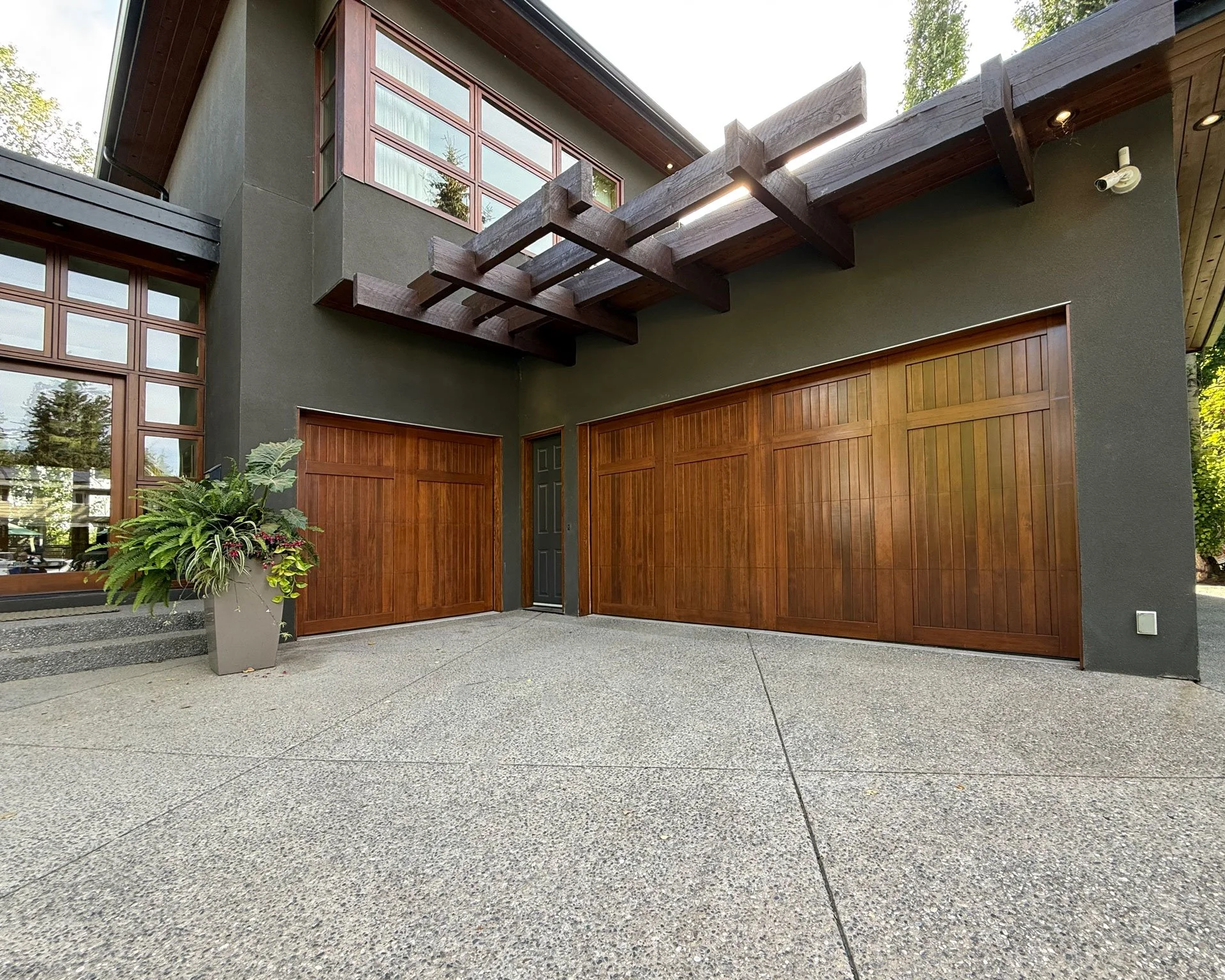A modern house with green exterior walls and wooden accents painted and stained by JC Paint, large windows, wooden garage doors, and a small front porch with a potted plant, outdoor lighting, and a security camera.