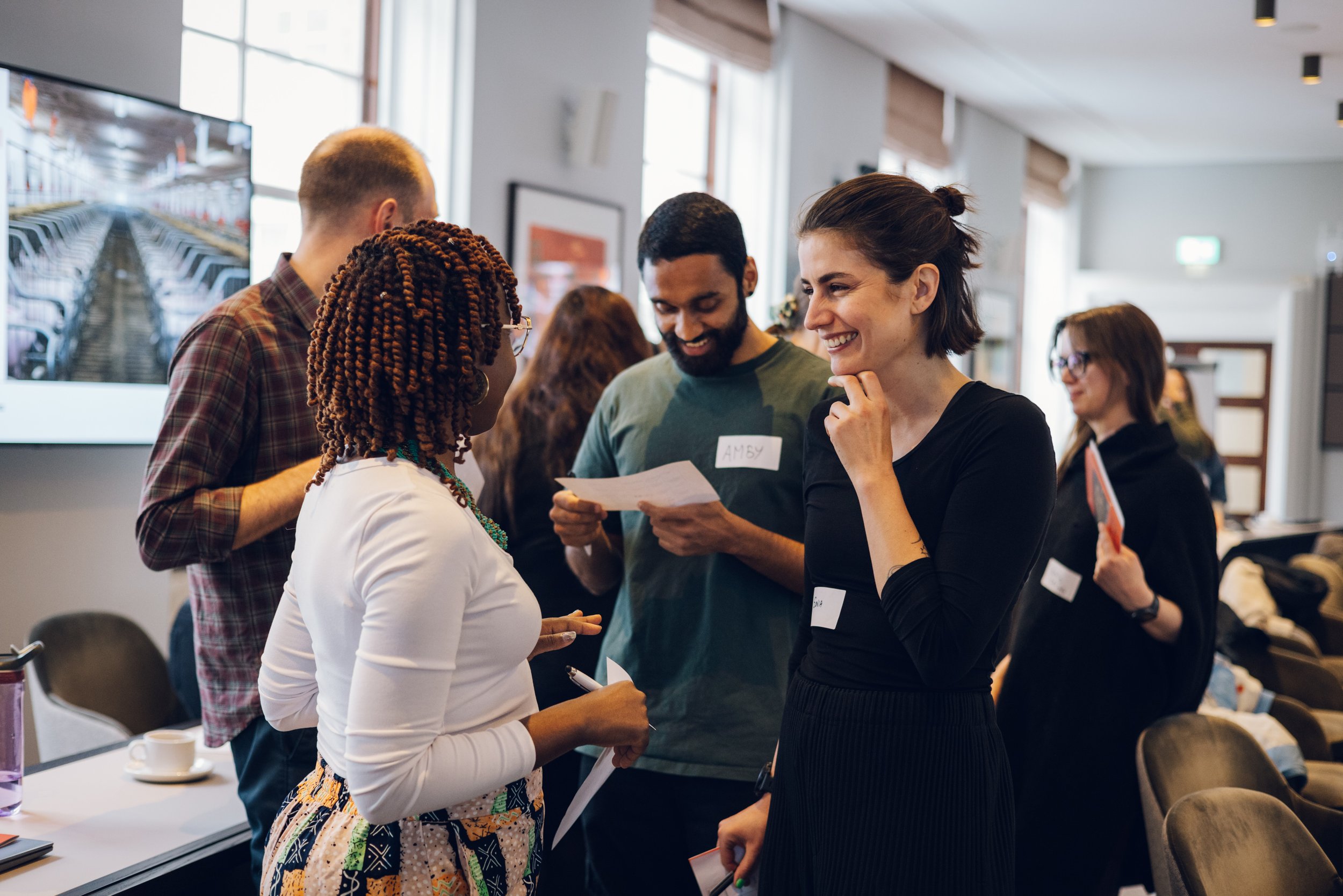 Group of people engaged in conversation at a professional or social event in an indoor conference room with large windows, some holding papers or taking notes, smiling and interacting with each other.