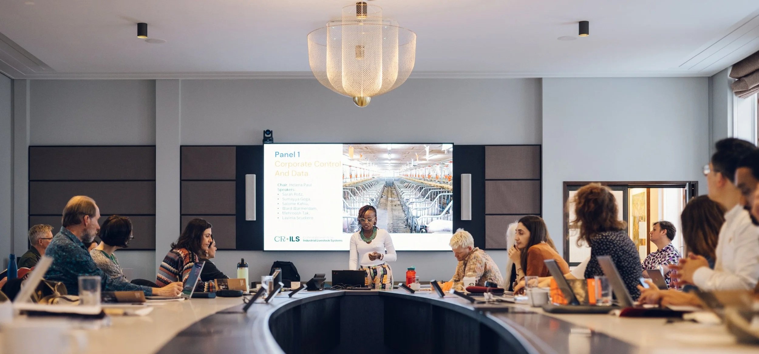 A diverse group of people attending a conference or meeting in a large room with a presenter at the front. The presenter is standing next to a screen displaying a presentation slide that says 'Panel 1: Corporate Control and Data.' The room has a large, curved table with laptops and notebooks, and a chandelier hanging from the ceiling.