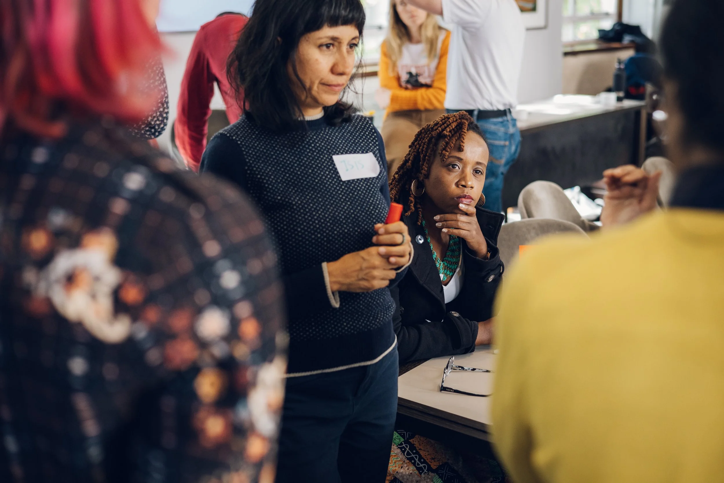 People standing in a circle at a workshop or seminar, with one woman in focus holding a red object, wearing a name tag, and others listening attentively.