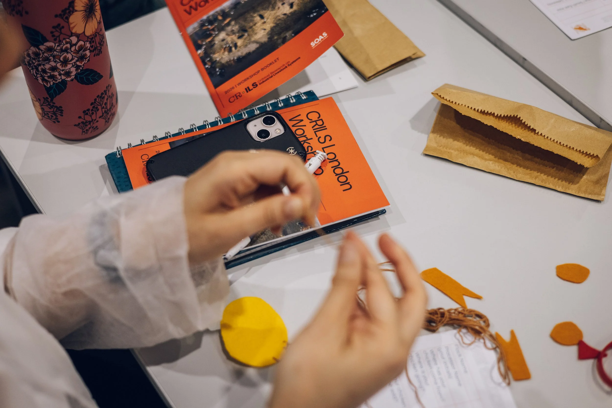 A person wearing a white long-sleeve shirt a spiral notebook, some yellow felt cutouts, paper, and yarn. The person is holding a small white object, a marker, and appears to be sewing.