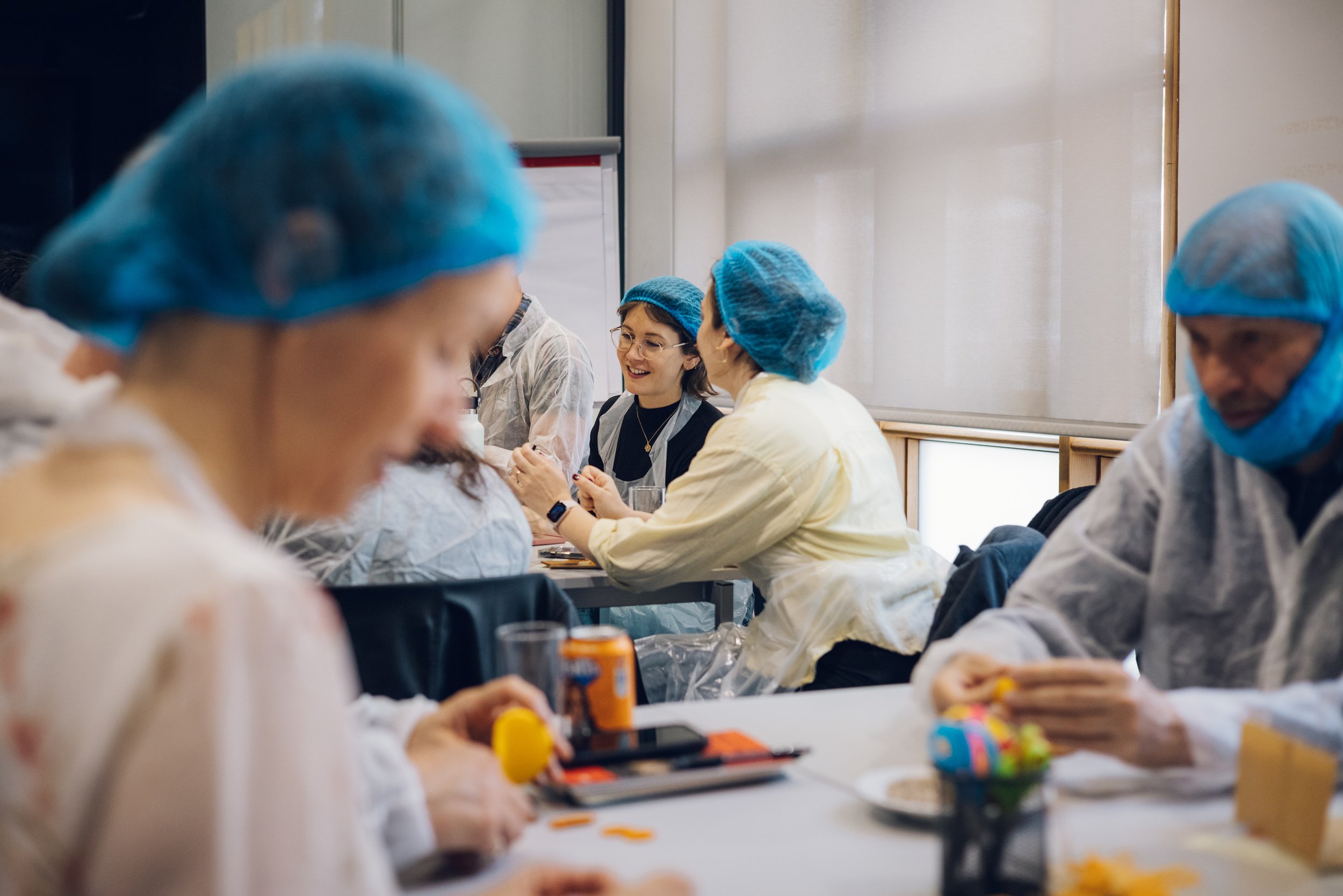 People wearing blue hairnets and white lab coats sitting at a table in a bright room. They are engaged in a creative activity, of arts and crafts, with various colorful materials on the table.